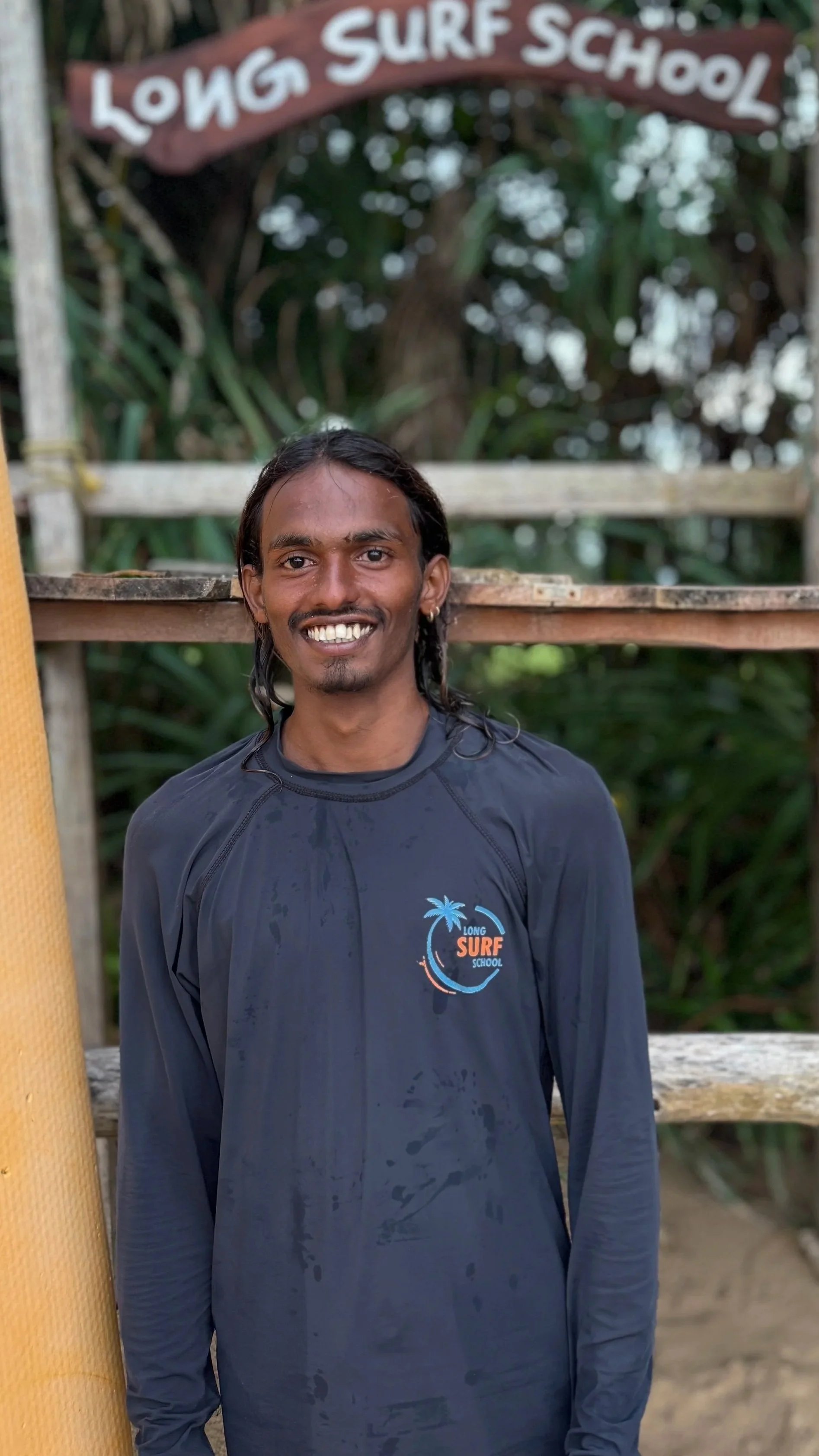 A man smiling for the camera, standing outdoors in front of a wooden structure with a sign reading "Long Surf School." He is wearing a dark long-sleeve shirt with a logo that says "Long Surf School" and features a palm tree and a wave. The background has green foliage and trees.