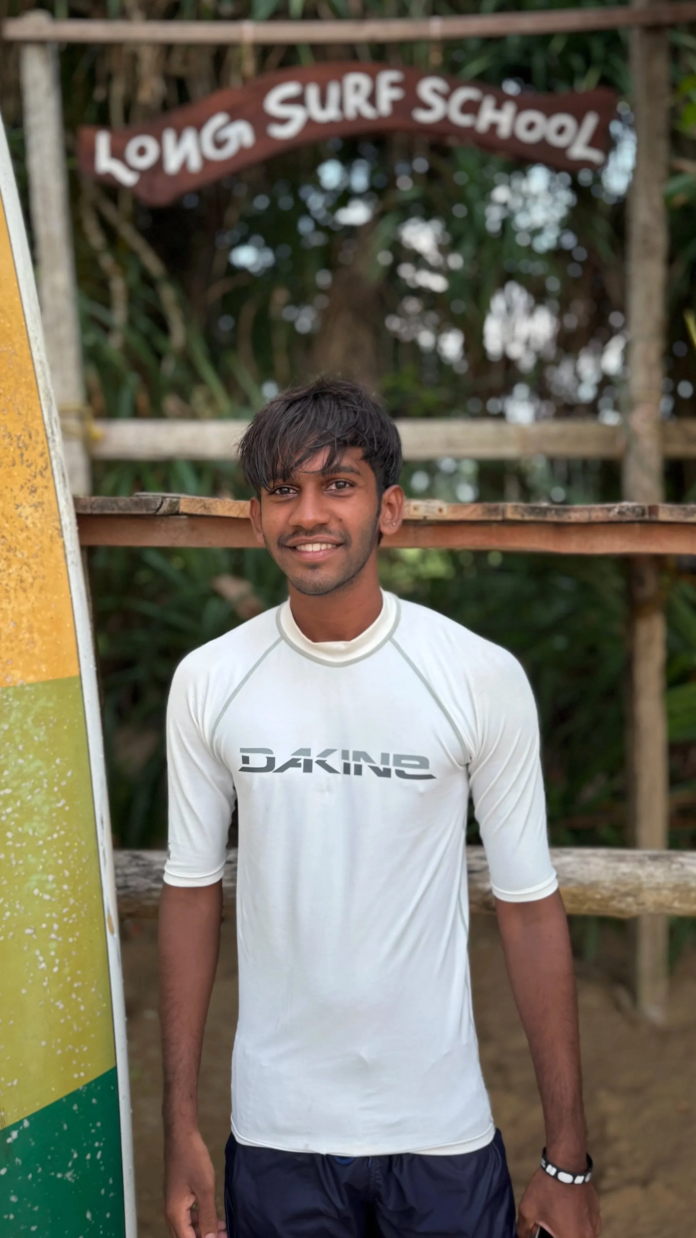 A young man wearing a white Daking sports shirt standing outdoors in front of a wooden surf school sign that reads "Long Surf School" with a background of trees.