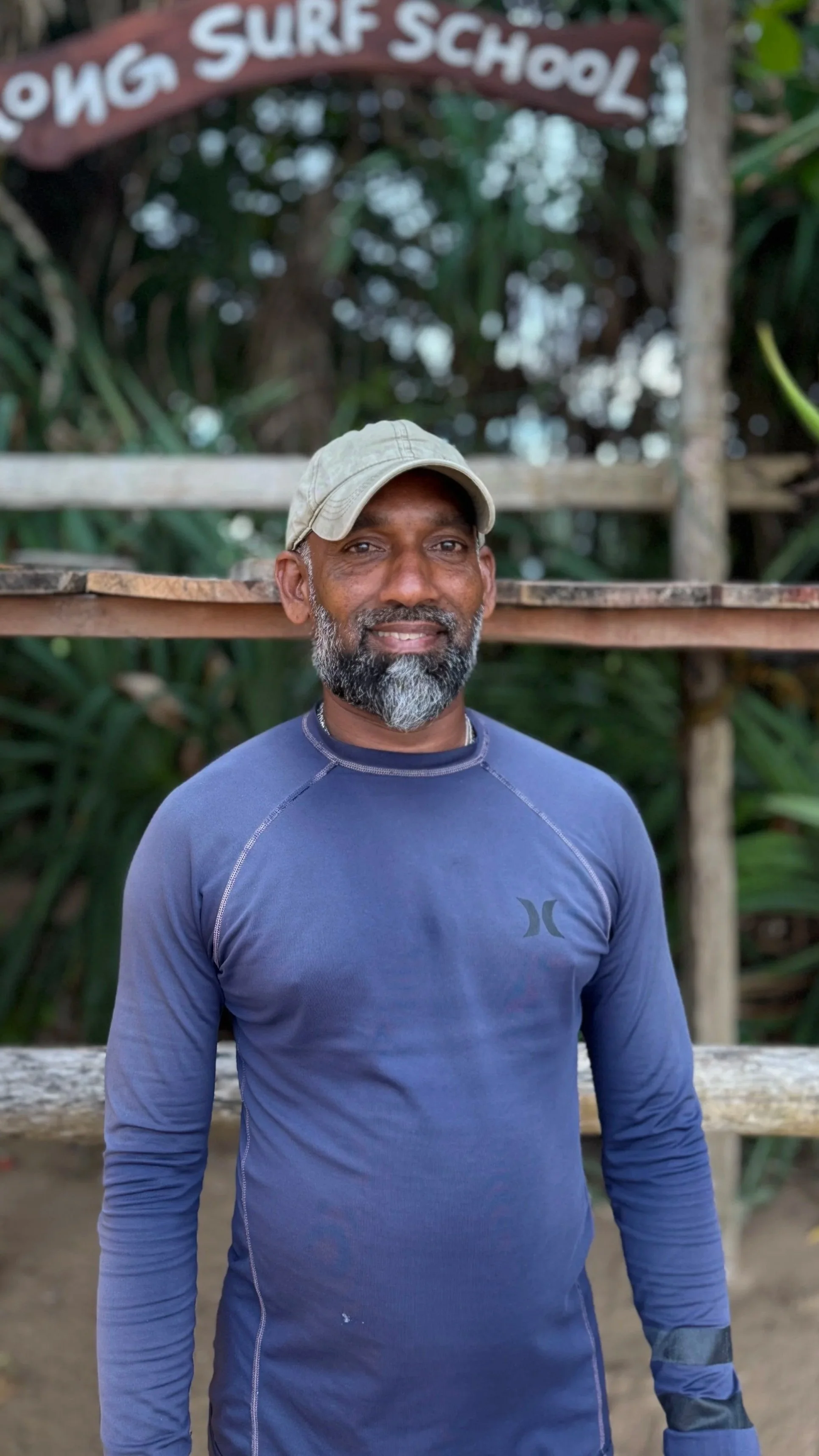 A smiling man with a beard wearing a beige cap and a blue long-sleeve athletic shirt standing outdoors with greenery and a wooden sign above his head that reads "Surf School."