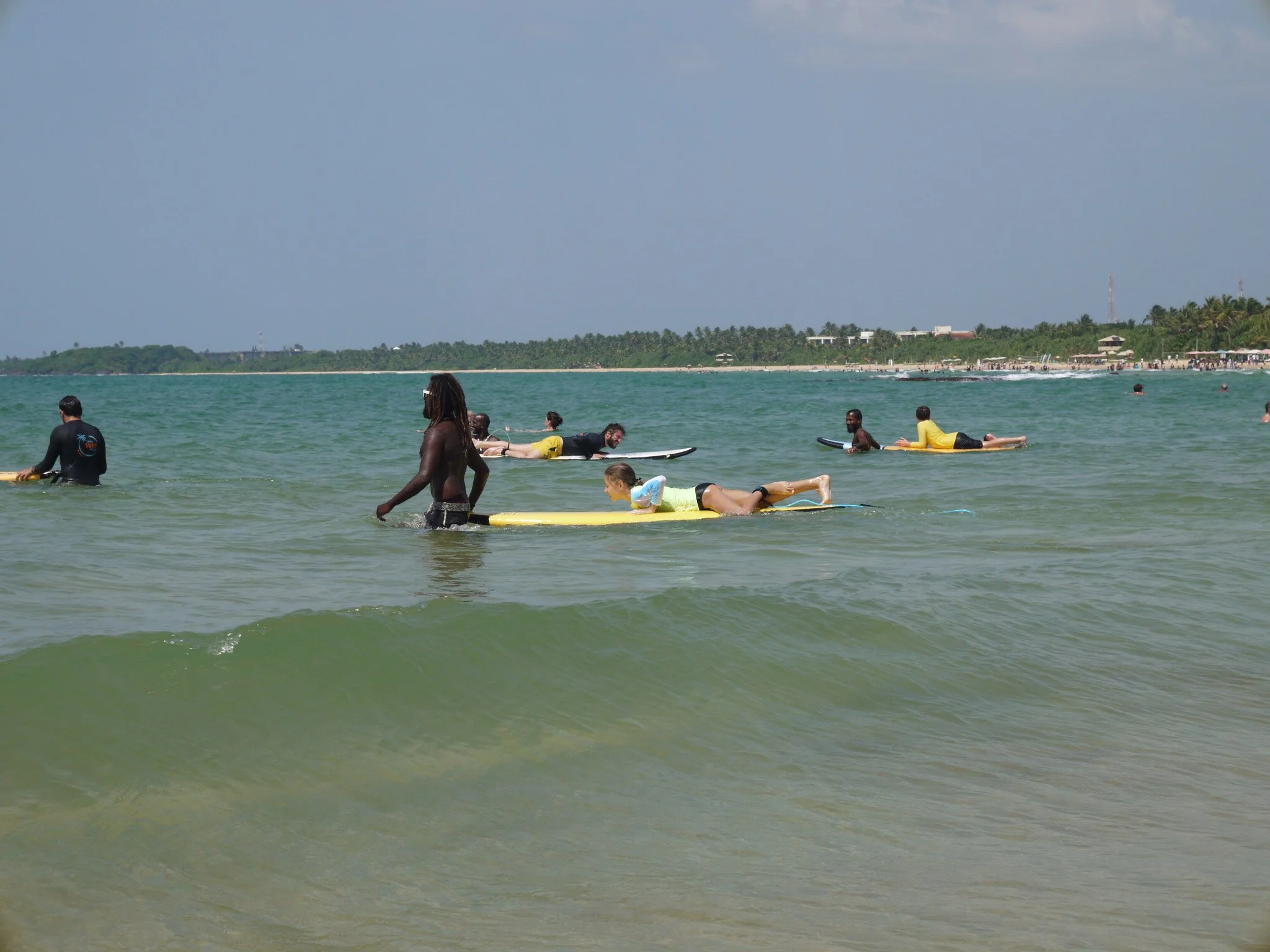 People surfing and swimming in the ocean near a beach with a forested coastline in the background.