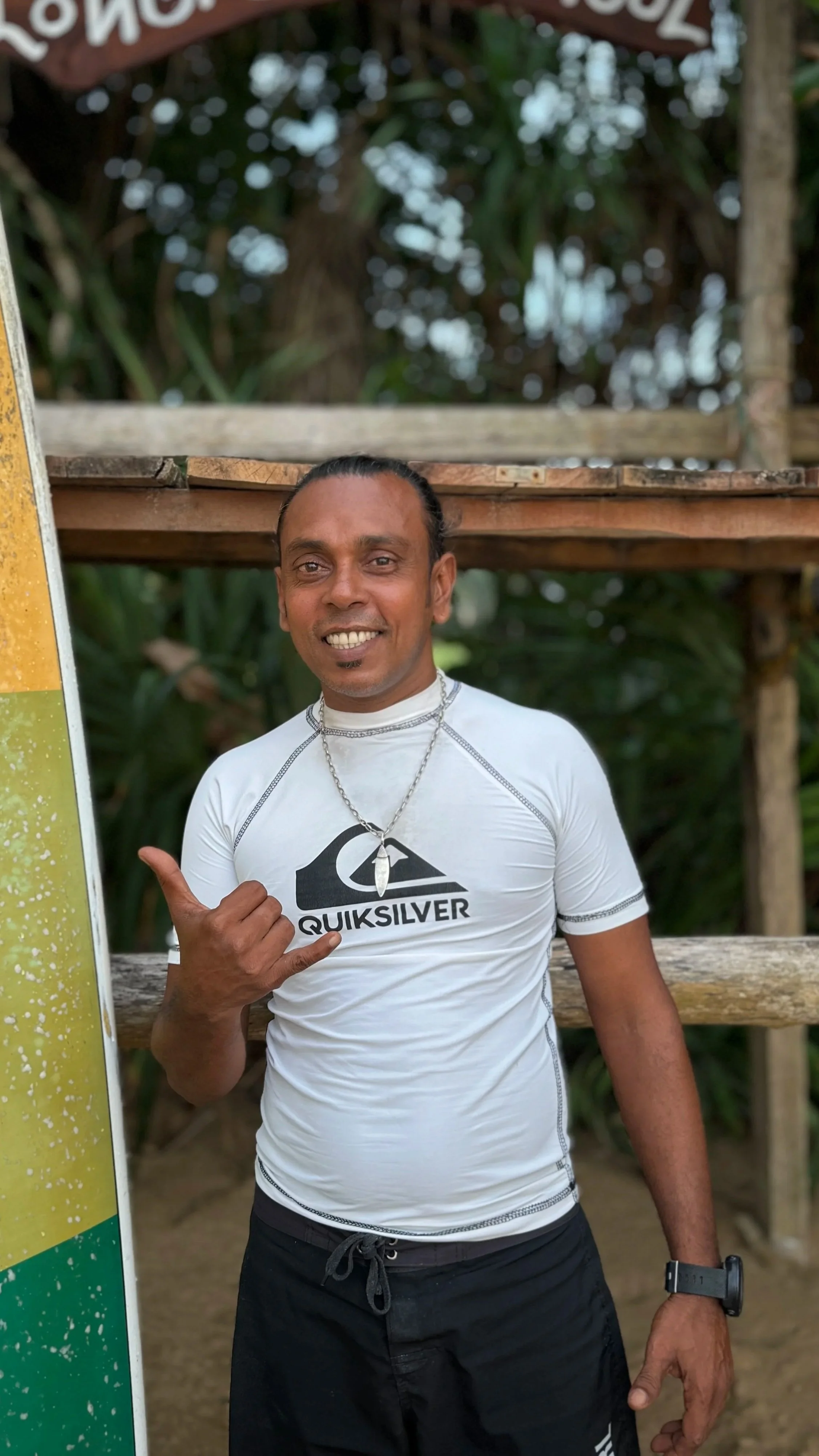 A man smiling and making a shaka sign with his right hand, wearing a white Quiksilver t-shirt, a black watch on his left wrist, and a necklace, standing outdoors with a wooden structure and greenery in the background.