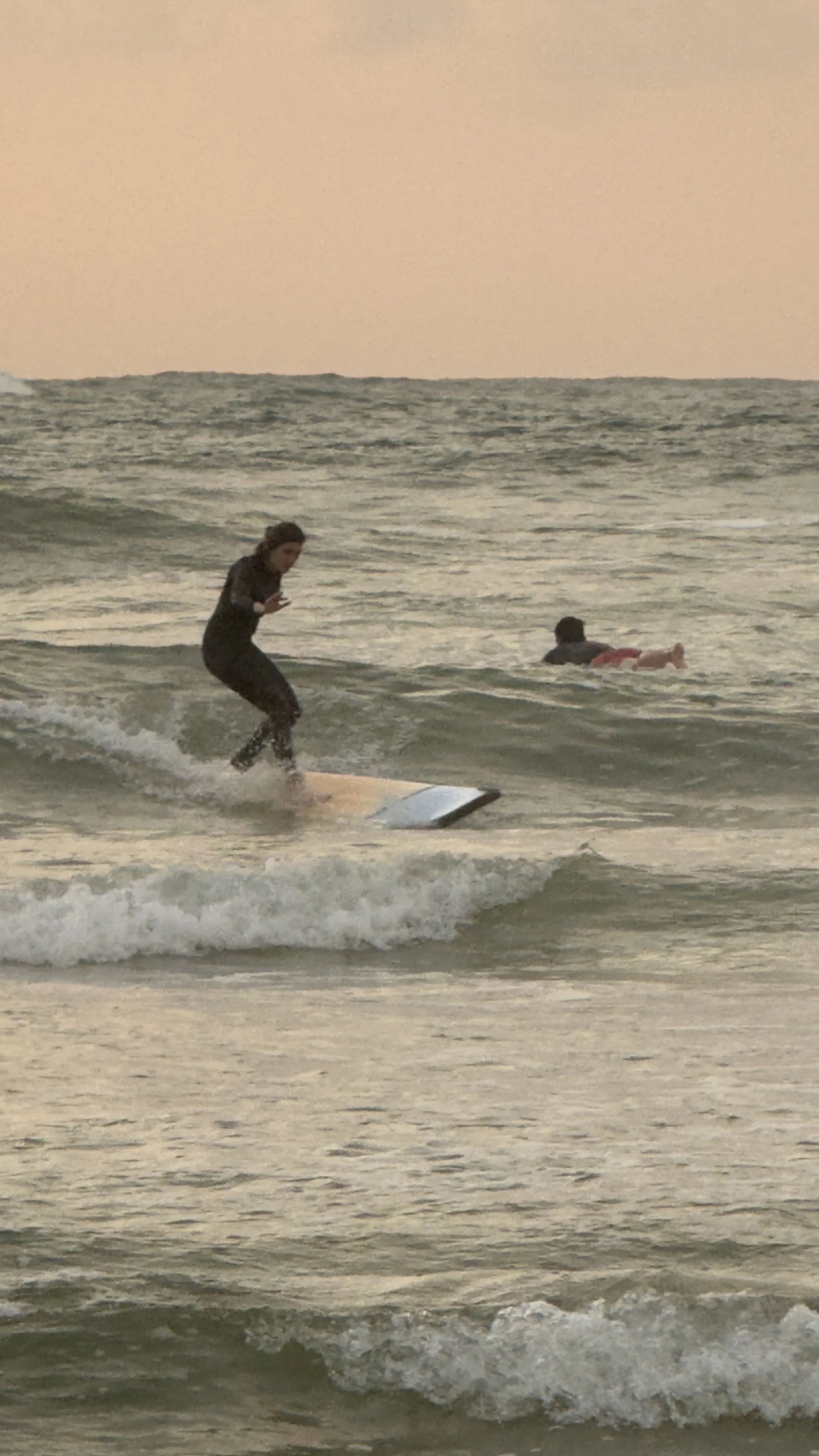 Person standing on a surfboard riding a wave in the ocean at sunset, with another person swimming in the background.