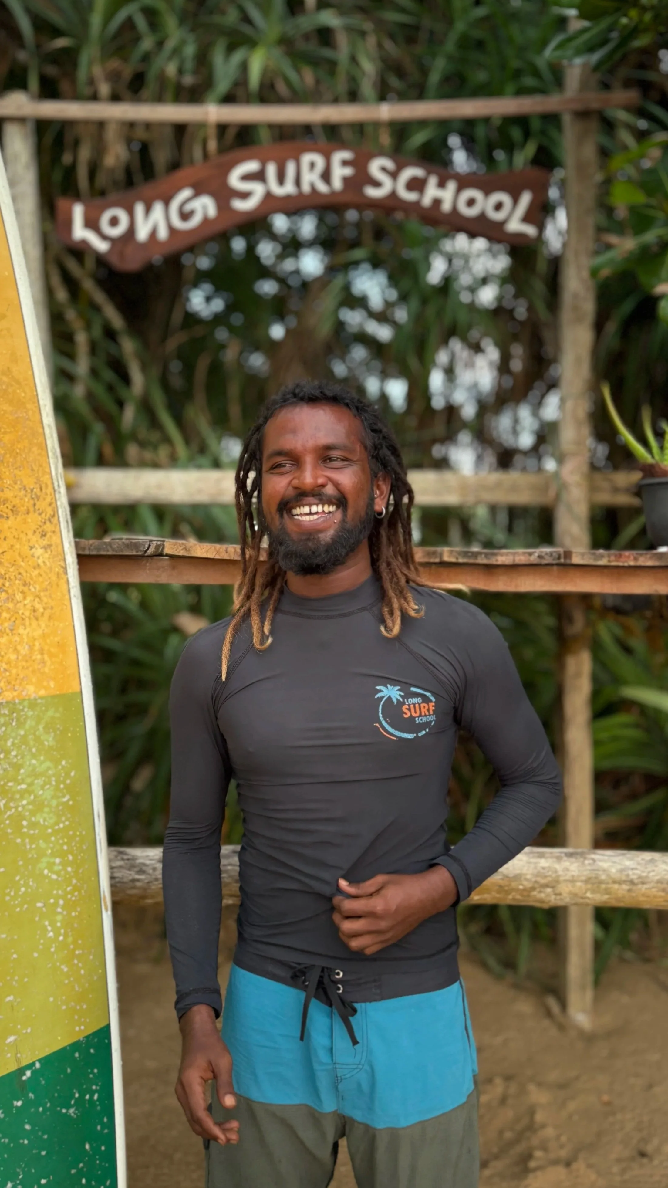 Smiling man with dreadlocks wearing a long sleeve surf school rash guard and blue swim shorts, standing in front of a sign reading 'Long Surf School' with greenery in the background.