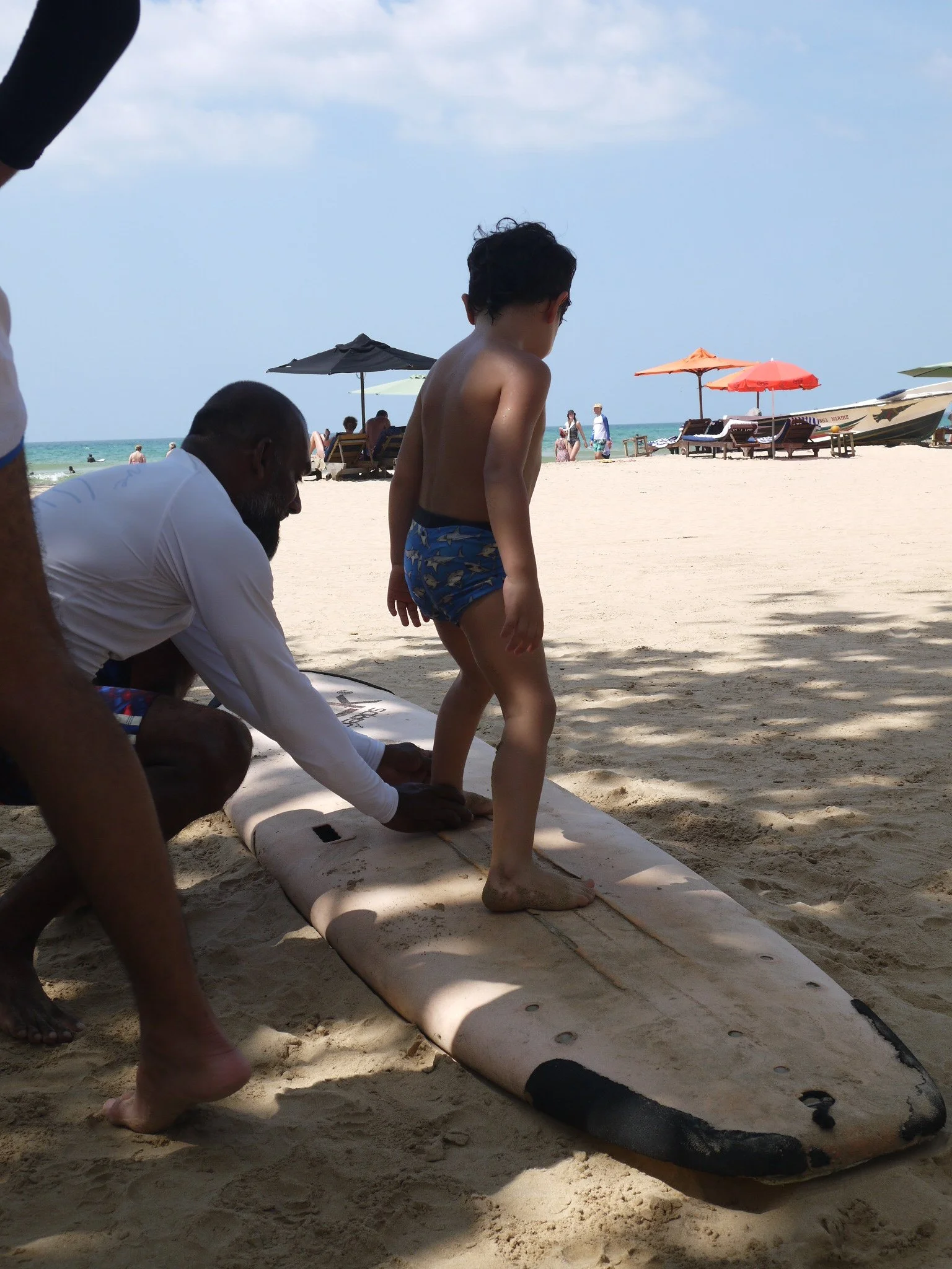 A boy in swim trunks stands on a surfboard on a sandy beach while a man kneels beside him, preparing to help or give instructions. The beach has umbrellas, lounge chairs, and other people in the background near the ocean under a partly cloudy sky.