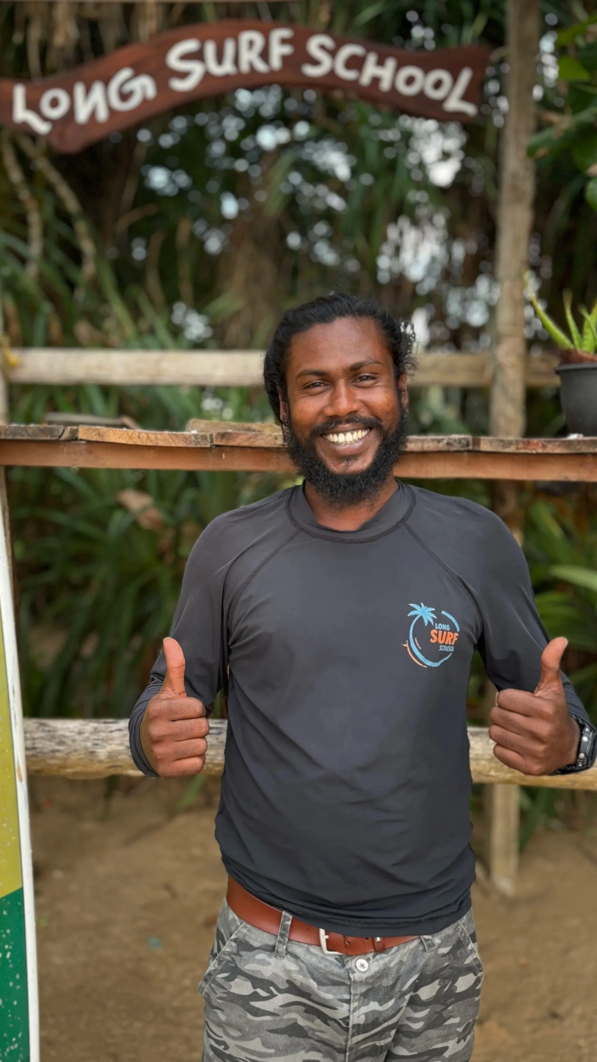 A smiling man with a beard giving two thumbs up in front of a sign that reads 'Long Surf School' surrounded by tropical greenery.