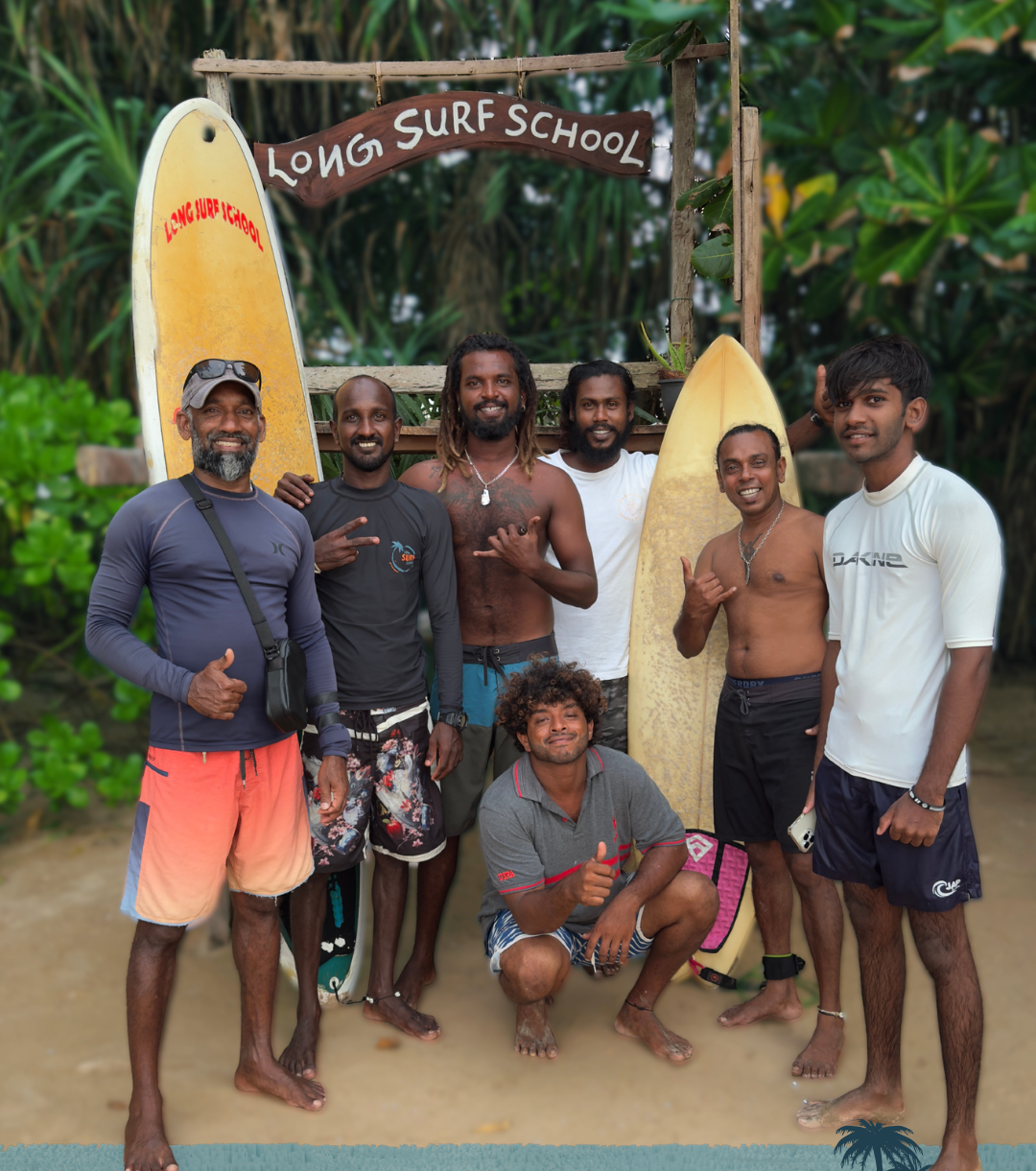 Group of six surfers standing on the beach in front of a wooden surf school sign and surfboards, surrounded by lush greenery.