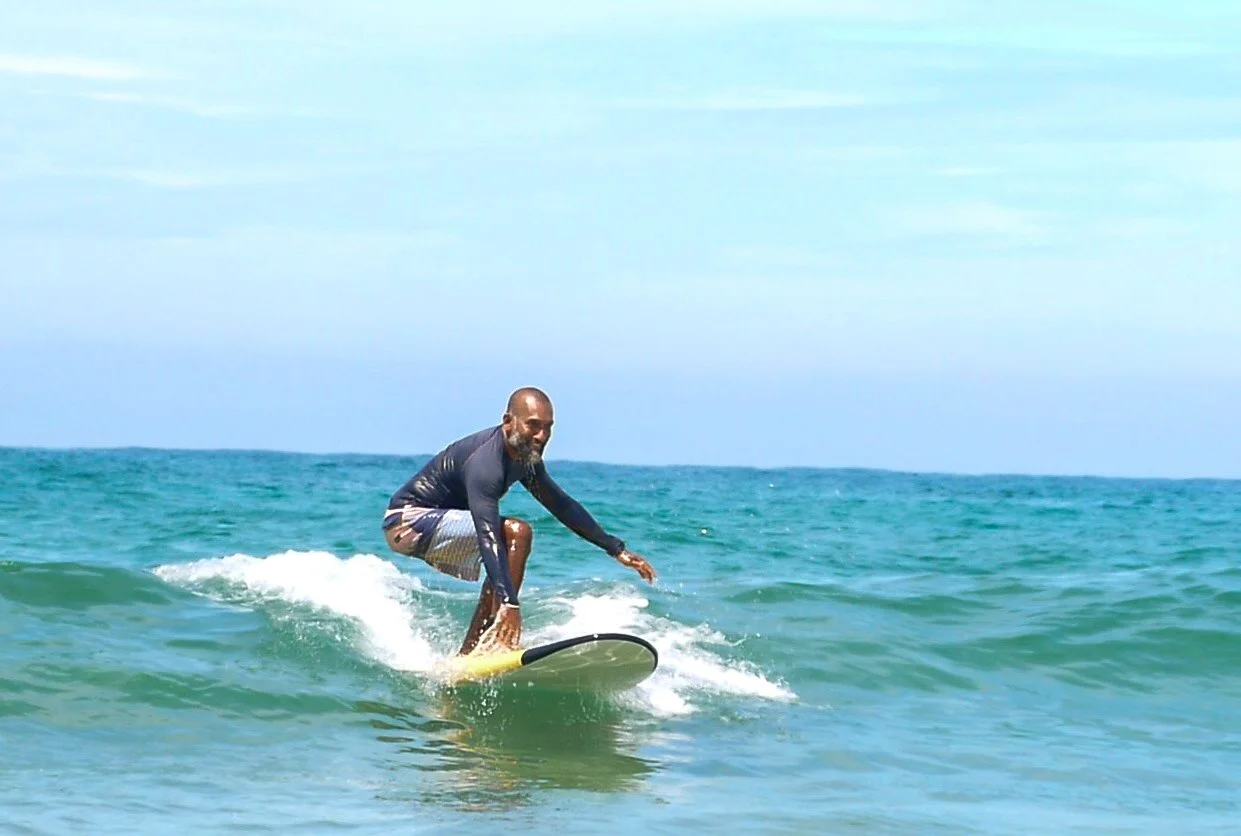 A man surfing on a yellow surfboard in the ocean, wearing a black long-sleeve shirt and dark shorts, smiling at the camera.