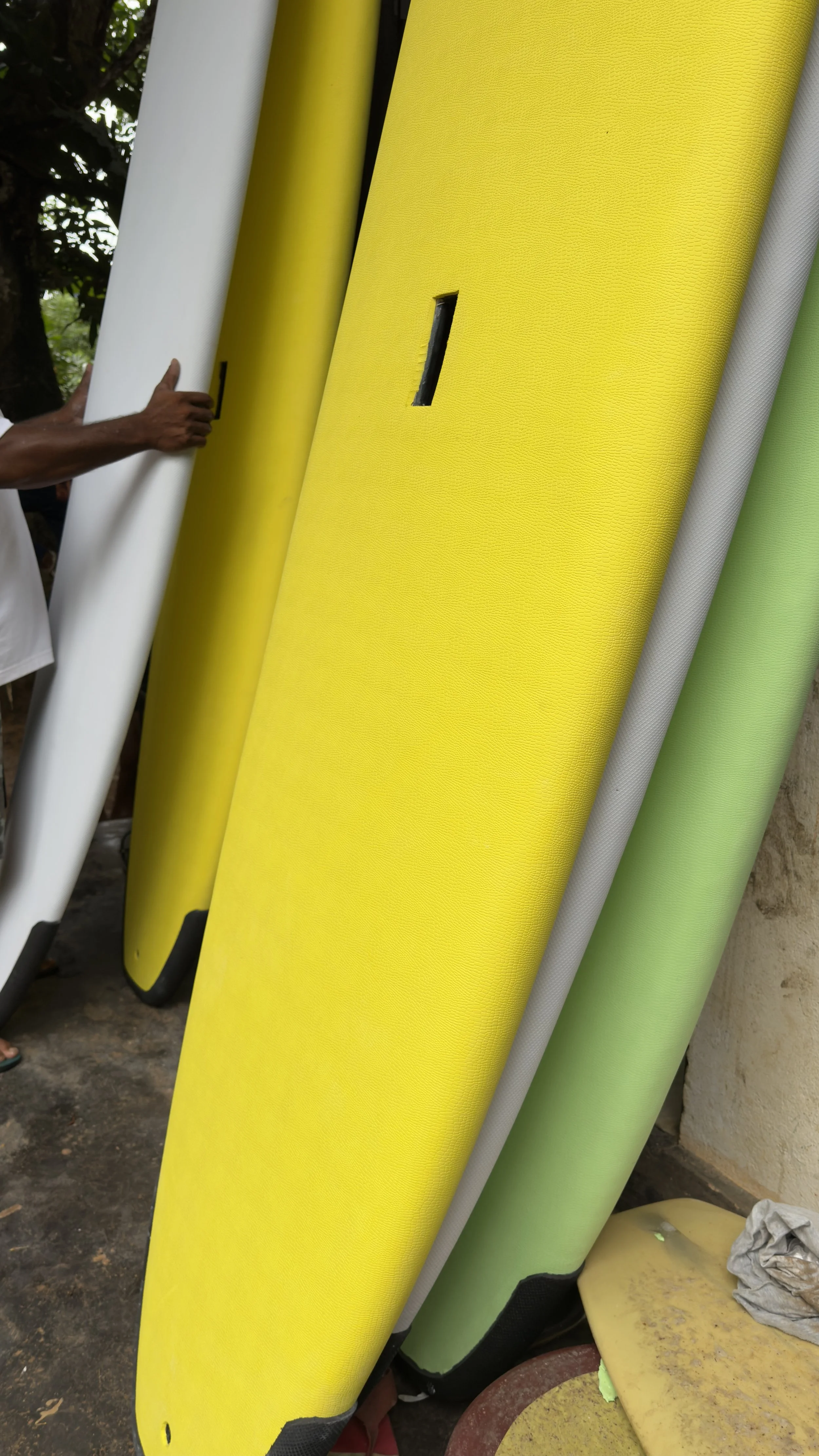 Several colorful surfboards leaning against a wall outdoors, with a person’s hand touching one of them.