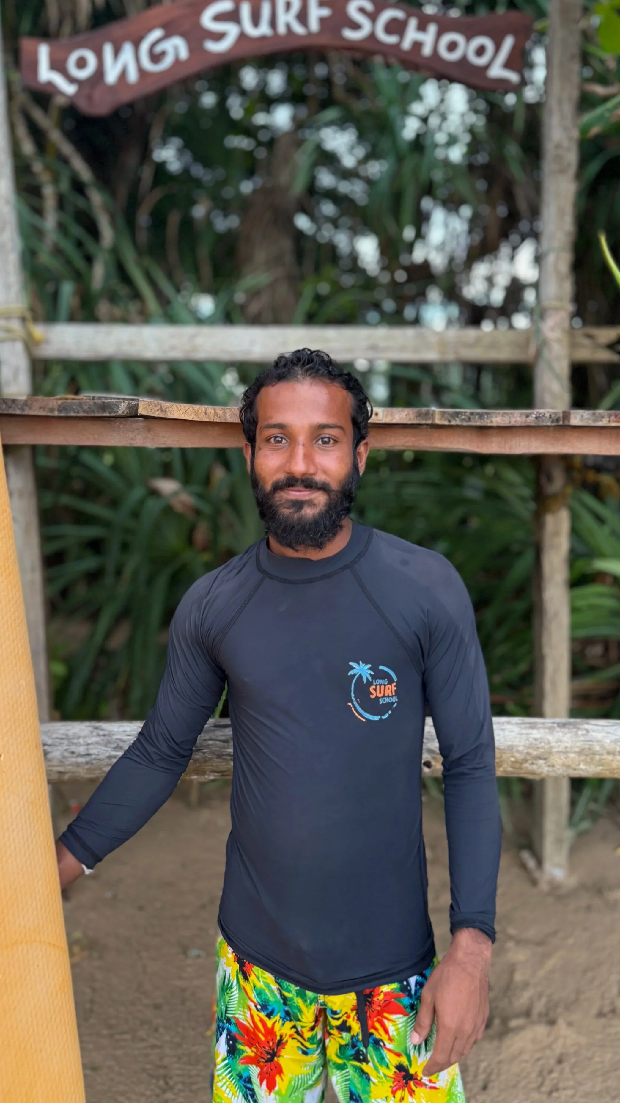 A man standing outdoors at a surf school, wearing a dark long-sleeve surf shirt with a logo, colorful tropical print shorts, and smiling at the camera. Behind him is a wooden structure with a sign reading 'Long Surf School' and lush greenery.