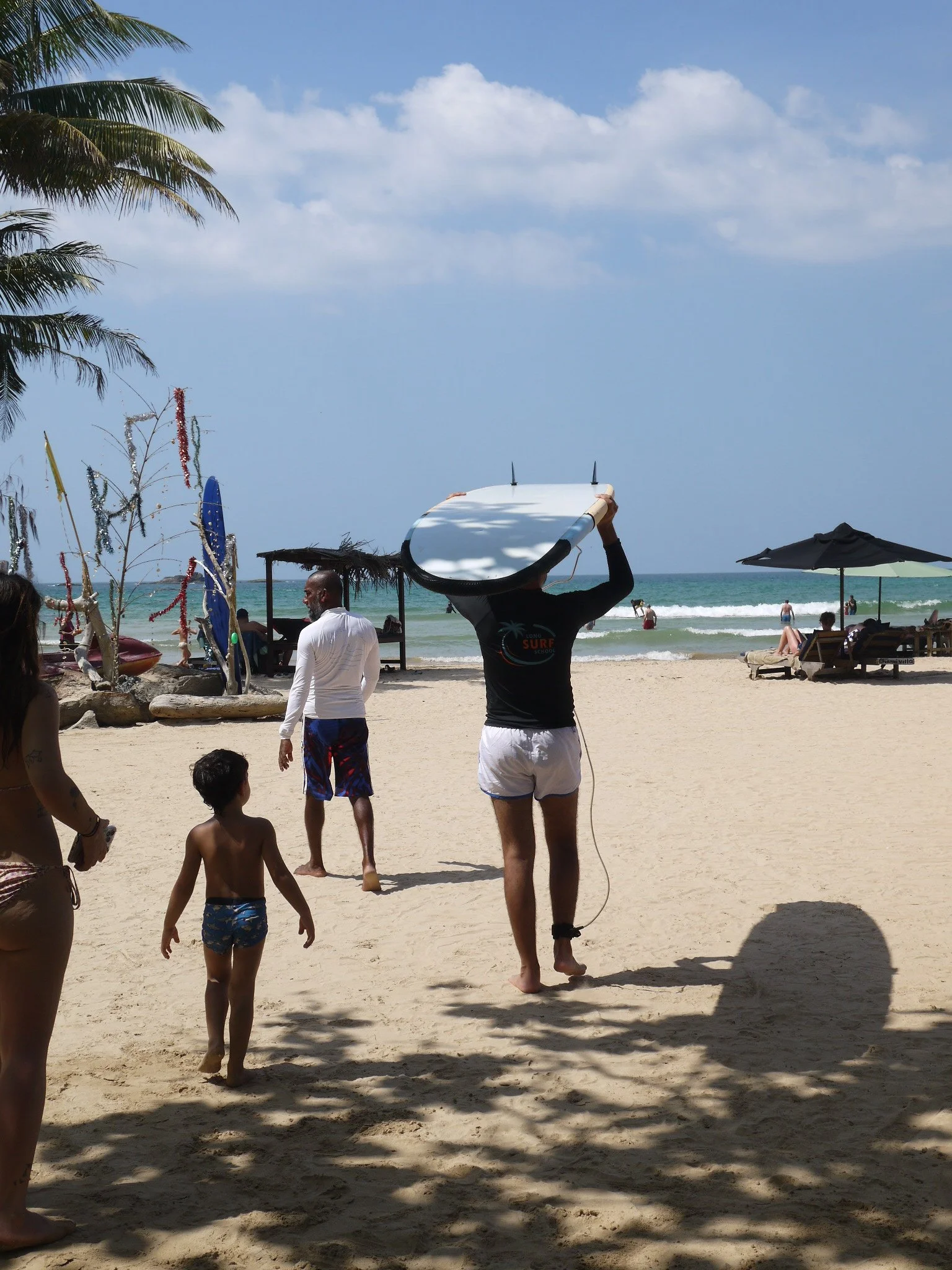 Beach scene with people, palm trees, umbrellas, and ocean, as someone carries a surfboard.