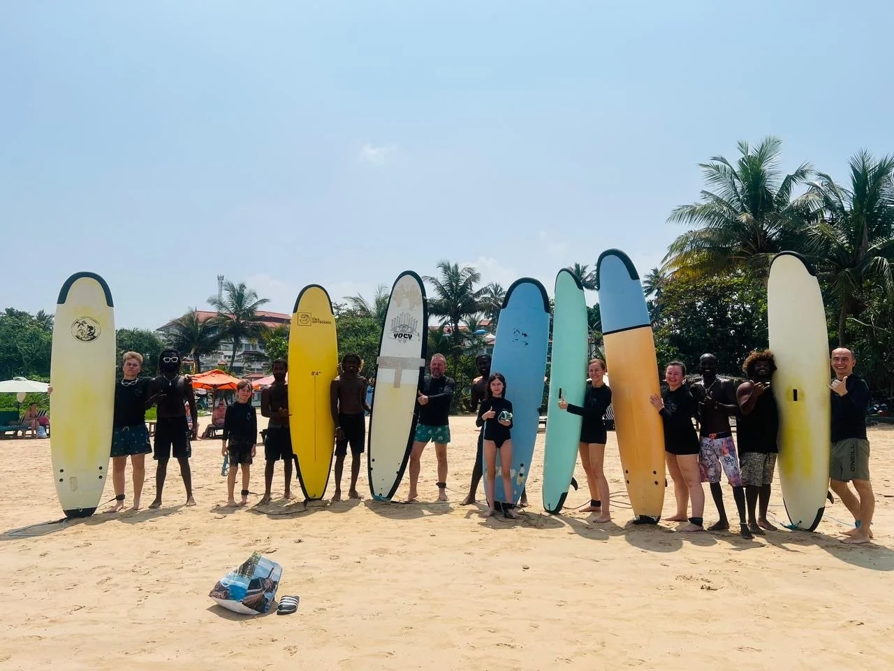 Group of people standing on a beach holding surfboards, palm trees in the background, sunny weather.