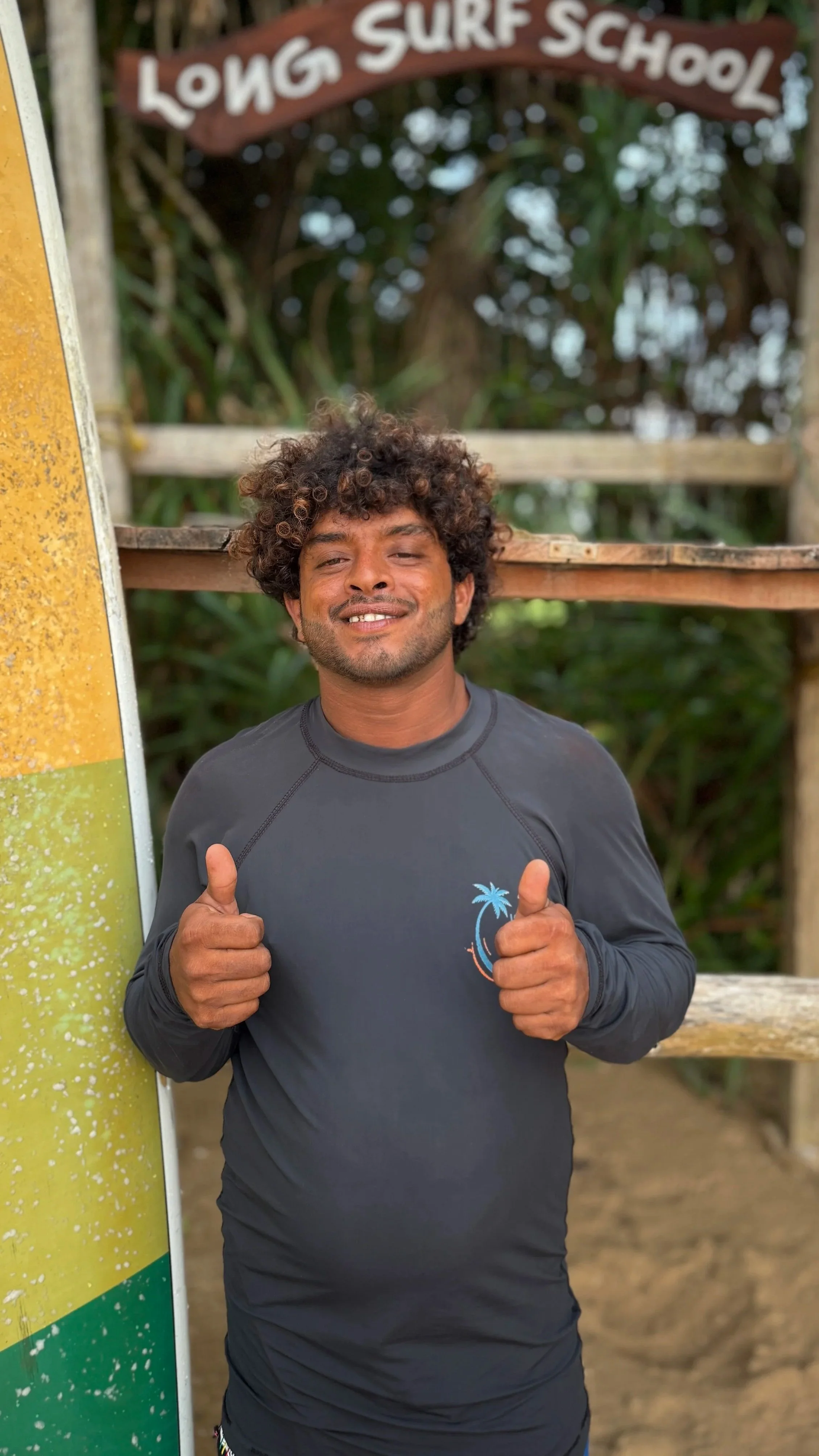 A man with curly hair and a beard giving two thumbs up, standing outdoors near surf school signage.