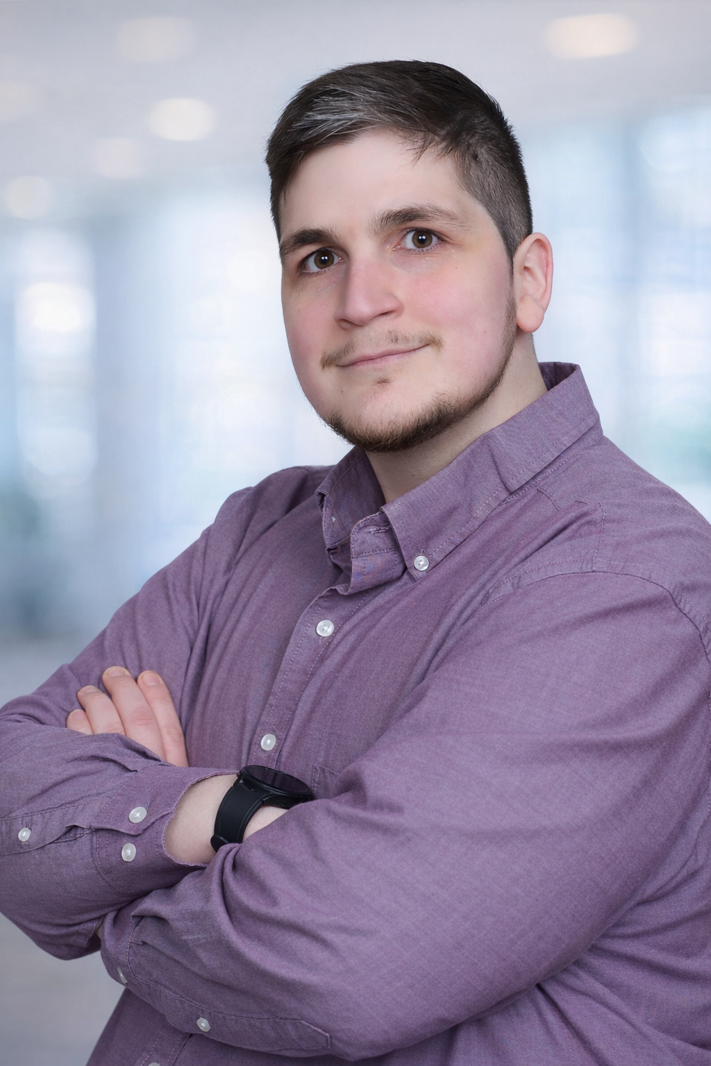 Portrait of a young man with short dark hair and a beard, wearing a purple button-up shirt, standing with arms crossed against a gray textured background.