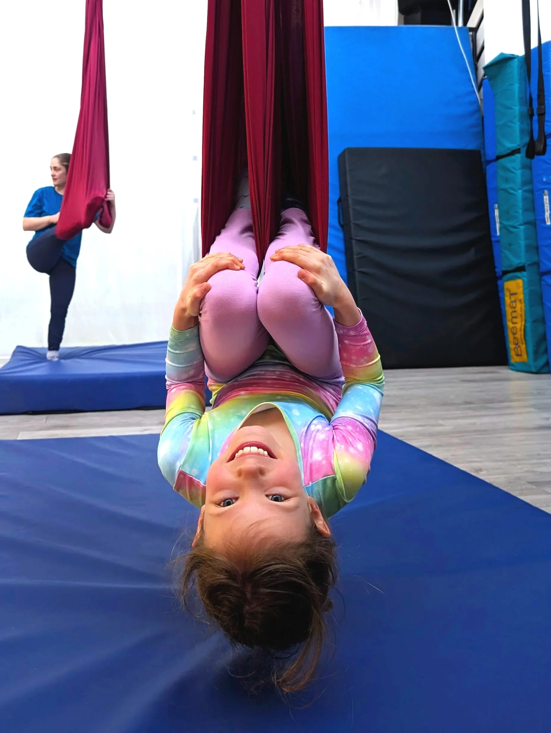 A young girl in a colorful tie-dye shirt and pink leggings lying upside down on a blue gymnastics mat, smiling at the camera. In the background, she is inside a fabric cocoon-like yoga or aerial silks setup, with another girl in a blue shirt practicing balancing on her leg on a raised mat.