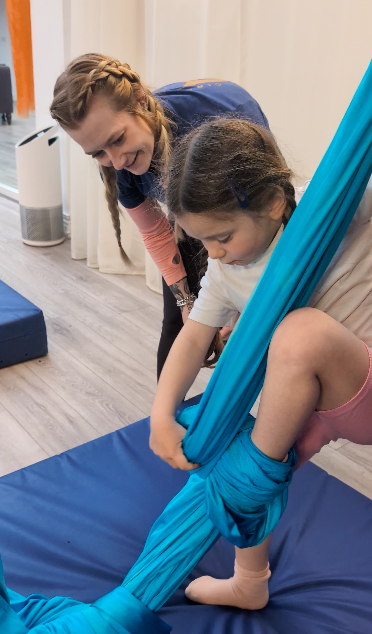 A young girl practicing aerial silk exercises while an instructor observes in a studio.