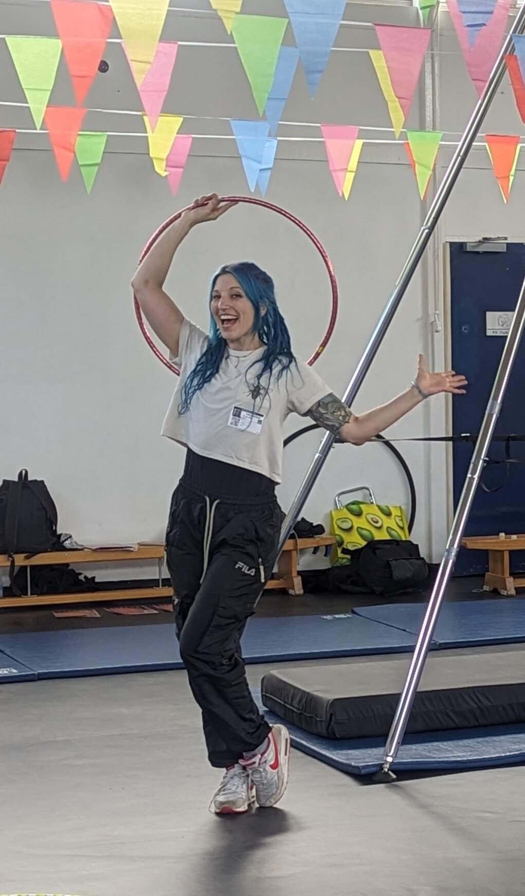 A young woman with blue hair smiling and balancing a hula hoop on her shoulder in a gym or circus training area, with colorful flags hanging from the ceiling.