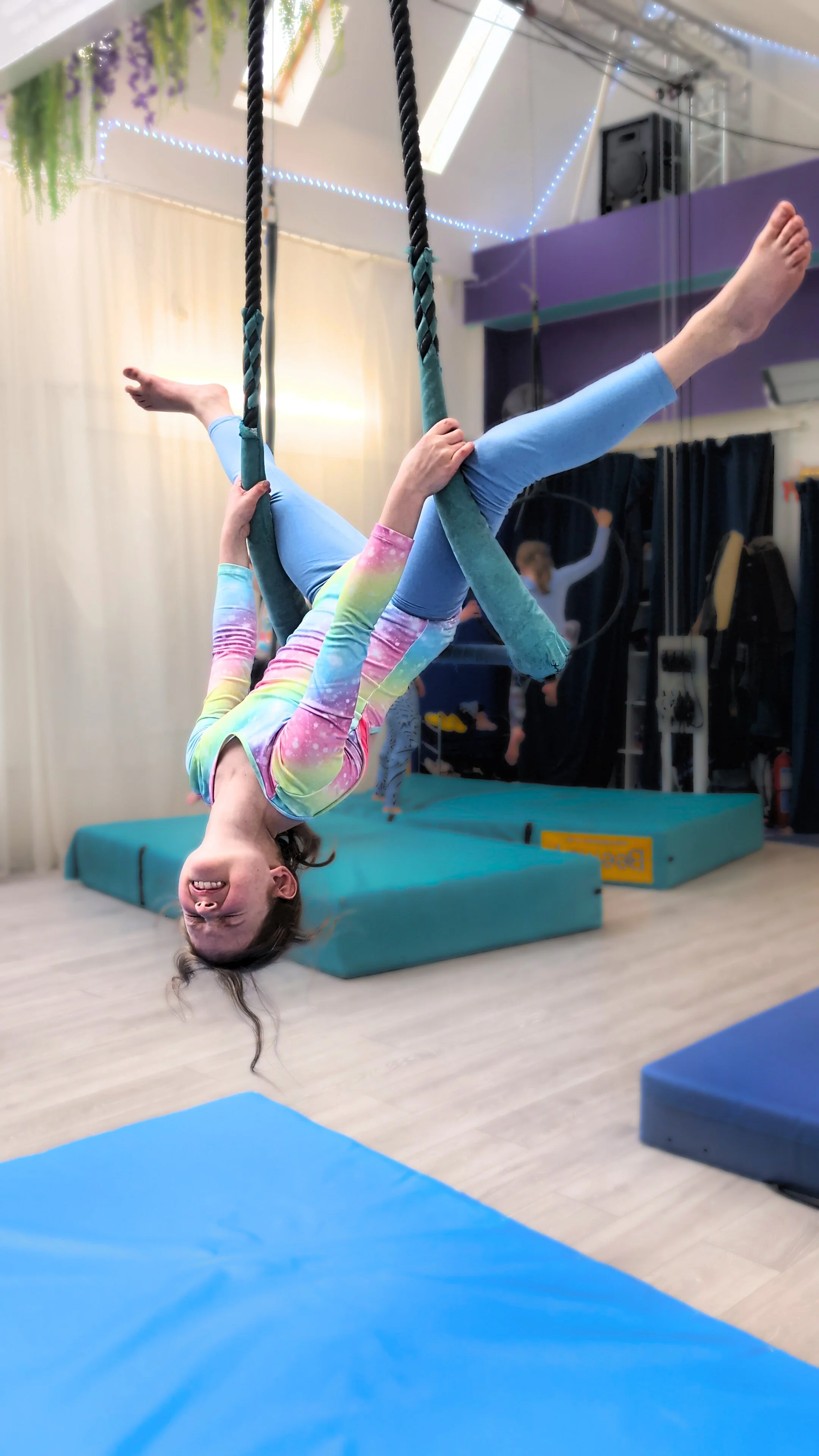 A young girl in colorful tie-dye clothing hanging upside down on a trapeze, in an aerial arts studio, smiling.