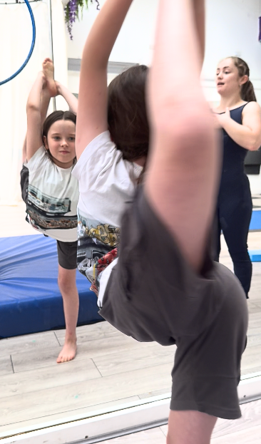 Children practicing gymnastics or dance in a studio, with a girl in a navy leotard and others stretching or posing.