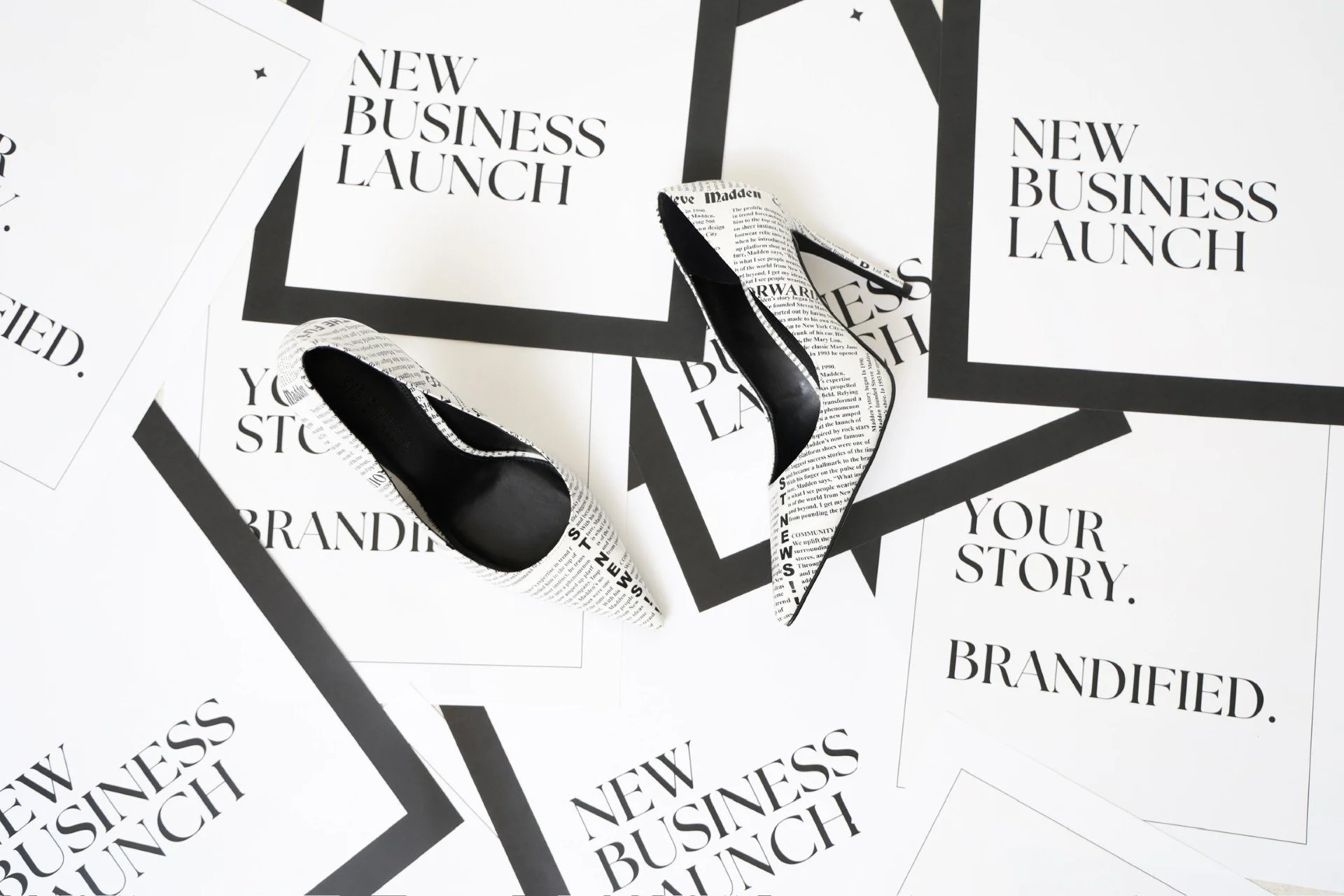 Pair of high-heeled shoes covered with newspaper print, placed on a surface with multiple flyers that read 'New Business Launch' and 'Your Story. Brandfied.' in black and white.