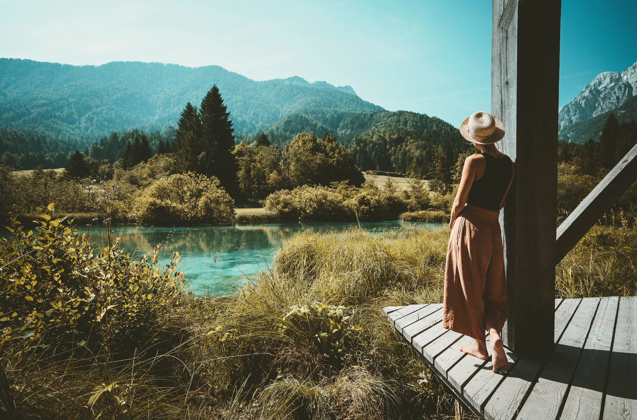 A woman in wide-brimmed hat and black top standing on a wooden deck, looking at a lake surrounded by trees and mountains.