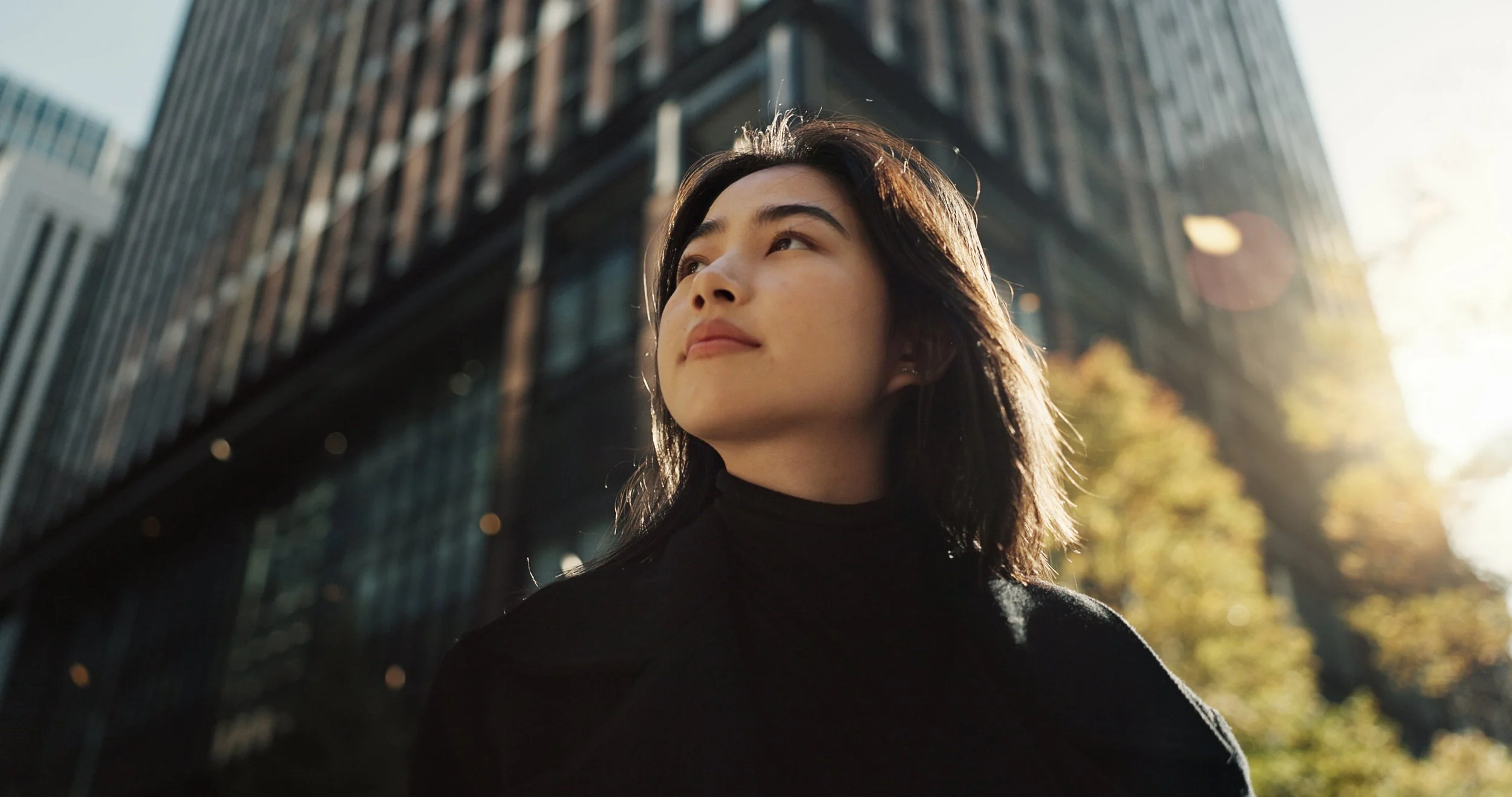 A woman with dark hair wearing a black turtleneck stands outdoors in a city, with tall buildings and trees with autumn leaves; sunlight shines from behind.