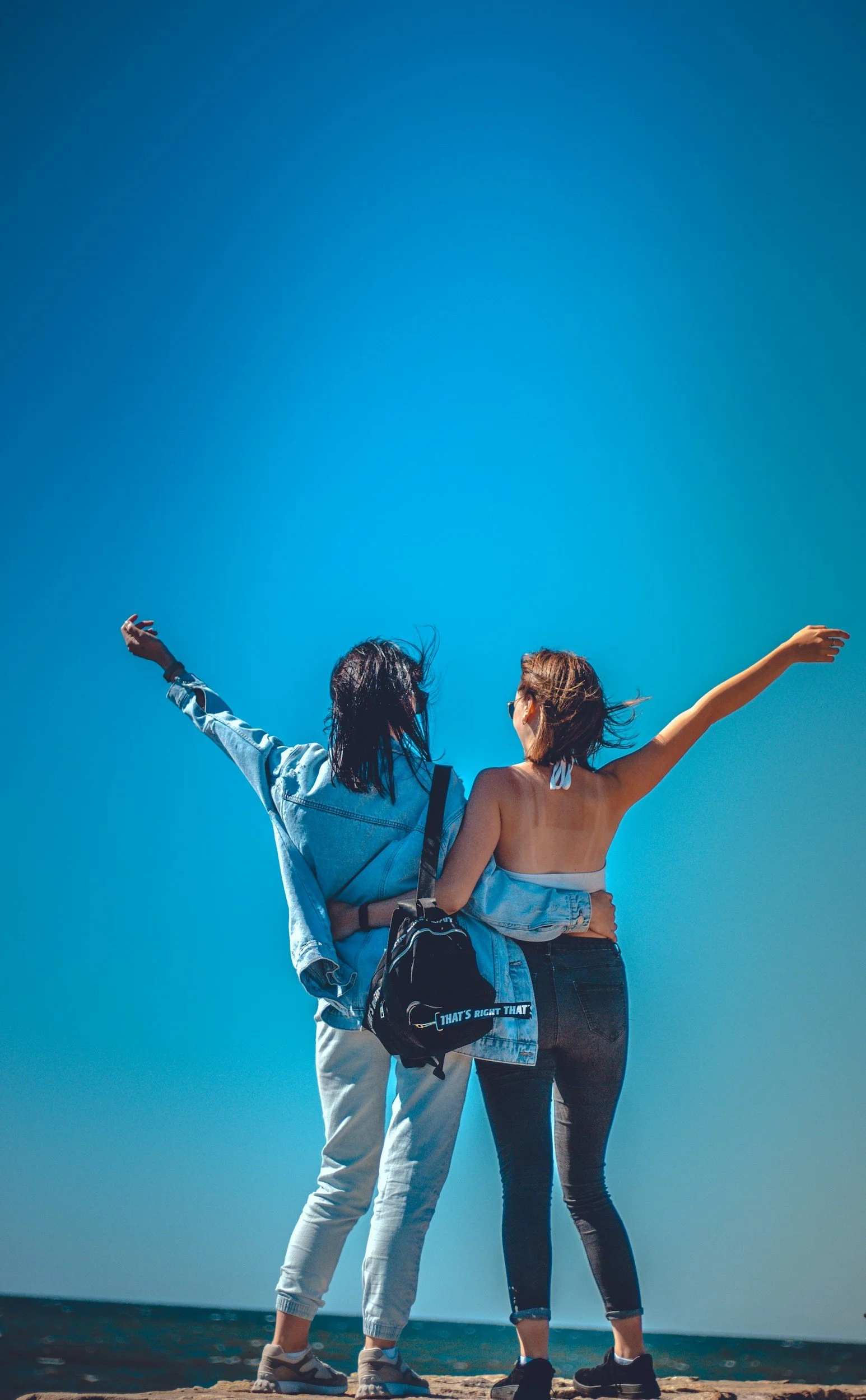 Two young women standing on the beach, facing away, with arms raised and hair blowing in the wind during daytime.