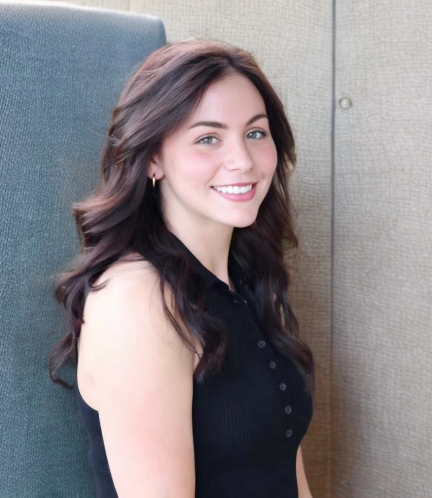 A young woman with long wavy brown hair, wearing a sleeveless black top, sitting and smiling indoors with a textured beige wall in the background.