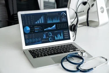 Laptop displaying data analytics and charts with a stethoscope on a white desk in a medical or office setting.