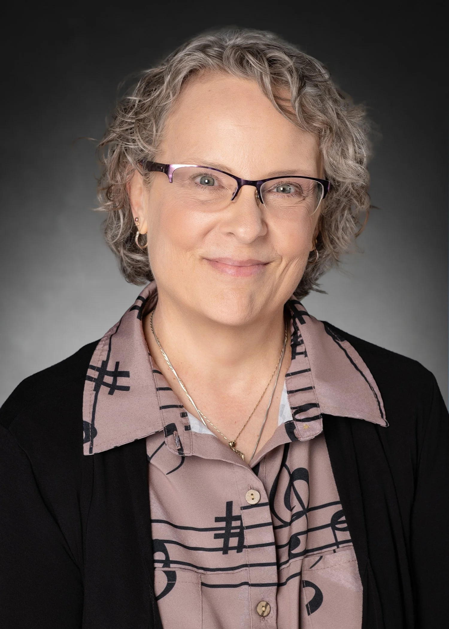 Close-up portrait of a woman with gray, curly hair wearing glasses, earrings, a patterned shirt, and a black blazer, smiling against a dark background.