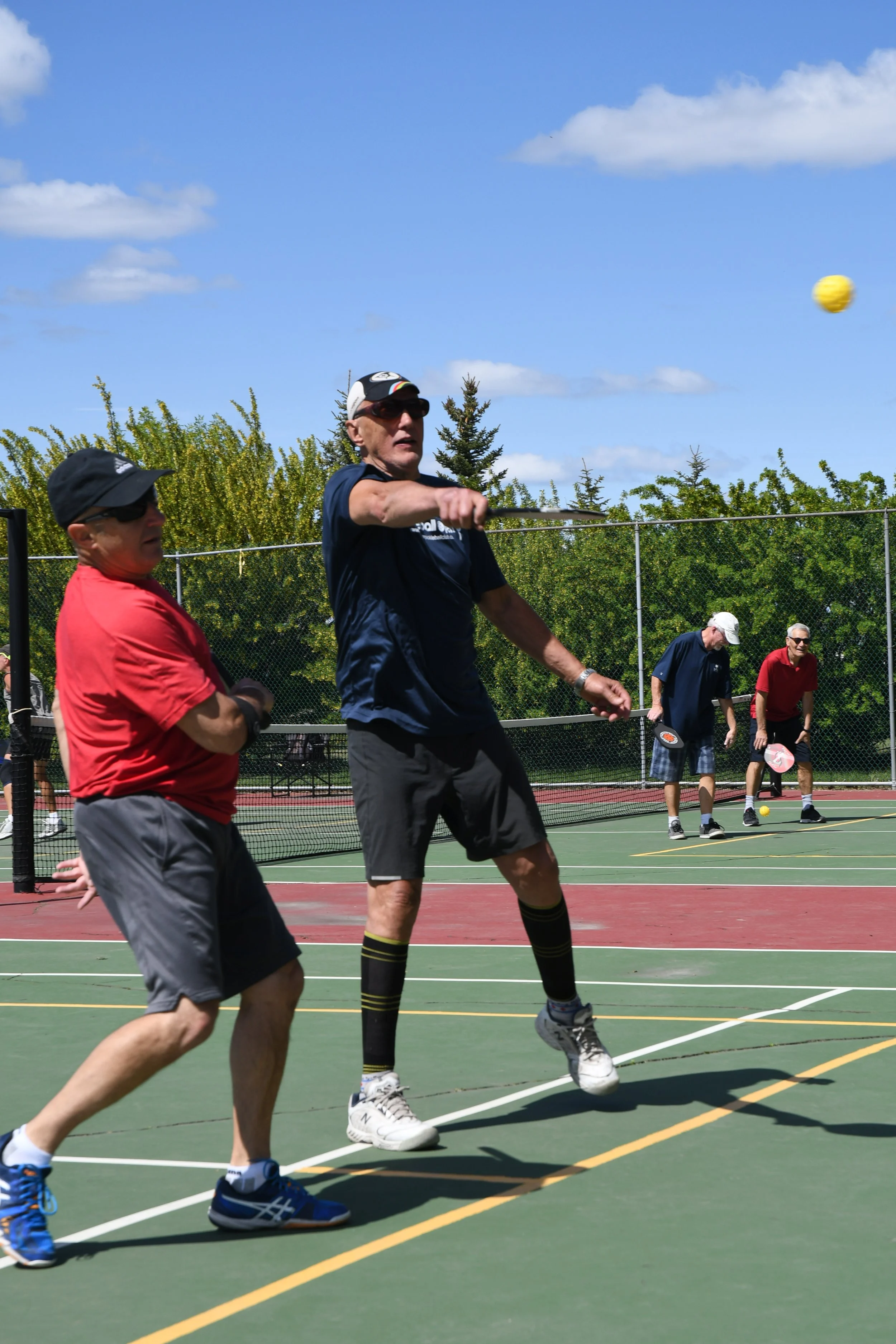 Older men playing pickleball on an outdoor court under a blue sky with clouds, some standing near the net and others in the background preparing for the next shot.