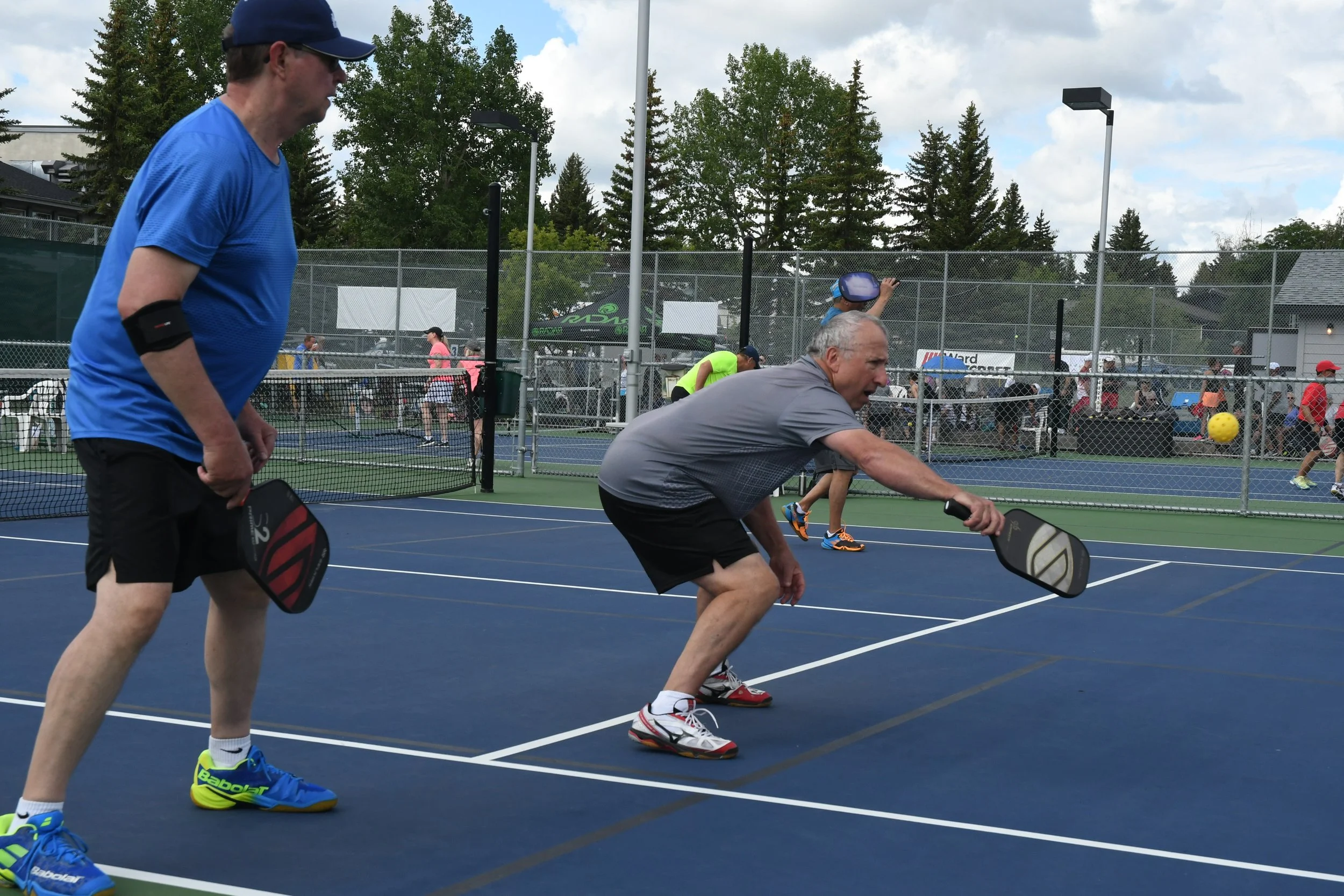 Two men play pickleball on a blue court, with one hitting the ball and the other watching, surrounded by a fenced area with people and trees in the background.