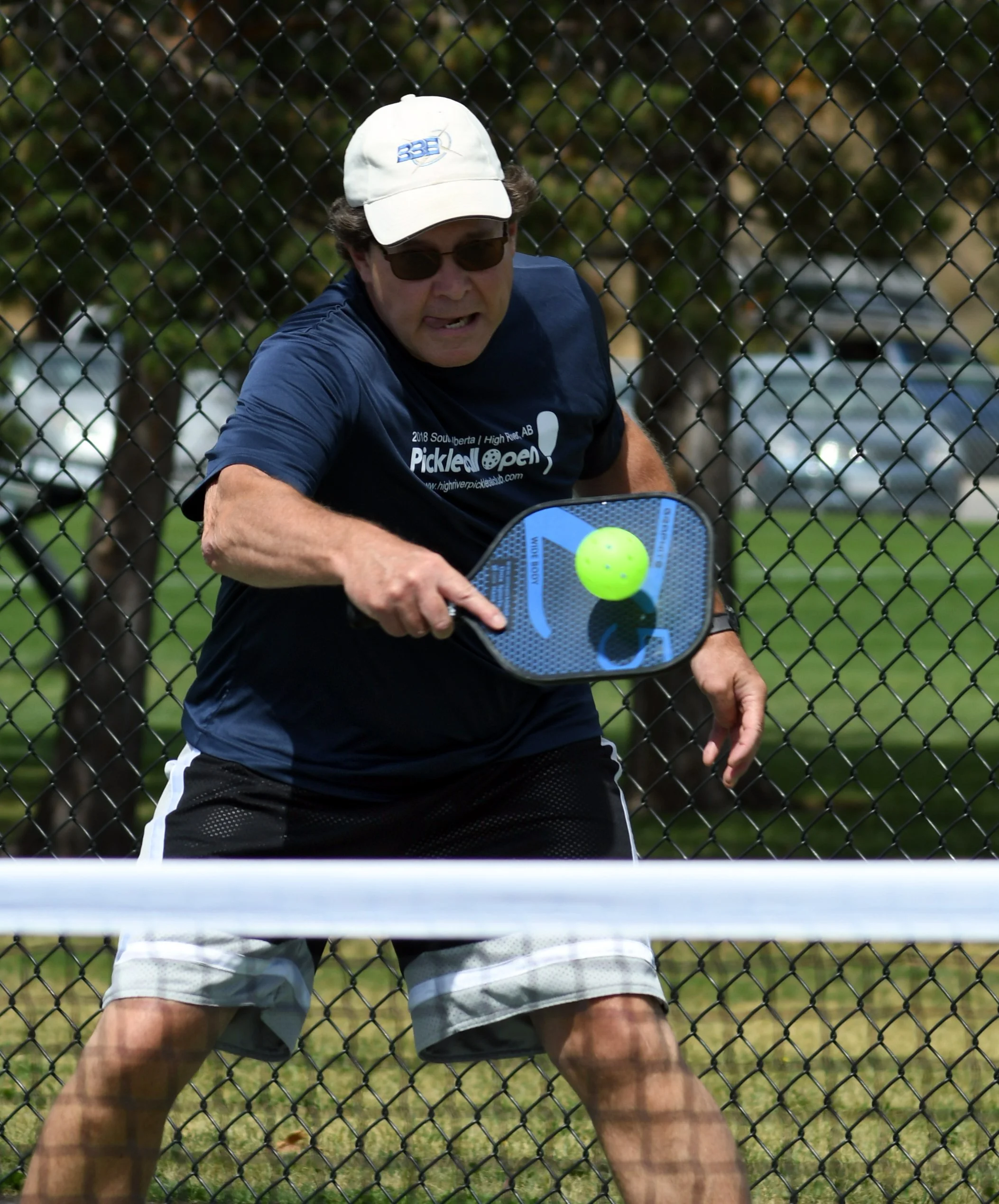 A man playing pickleball on an outdoor court, hitting a bright green pickleball with a paddle, behind a chain-link fence.