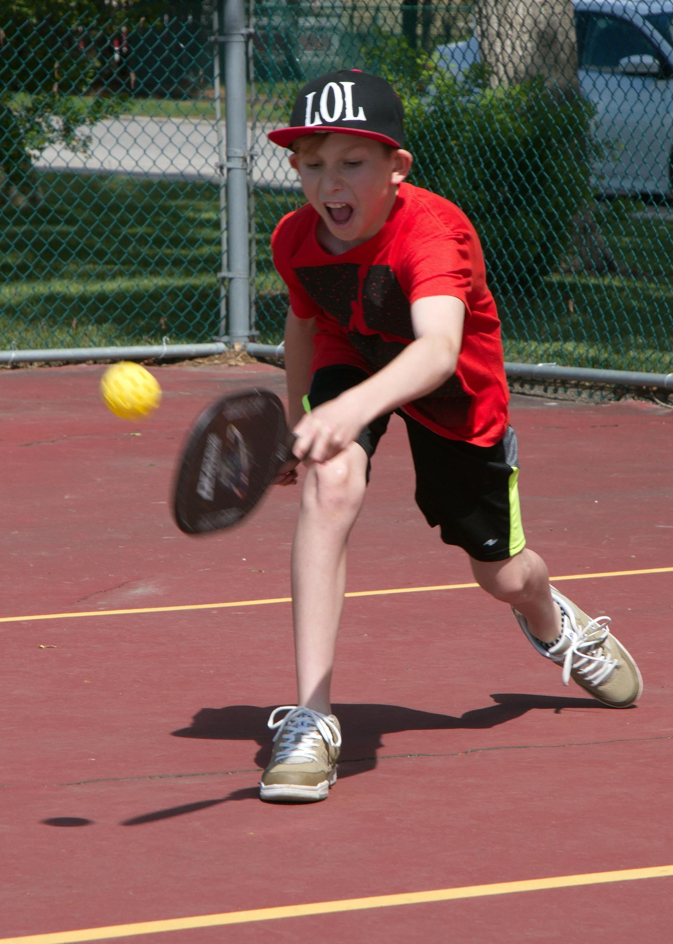A young boy wearing a black cap with 'LOL' in white letters, a red t-shirt, black shorts with neon green accents, and beige sneakers playing pickleball on an outdoor court, hitting a yellow ball with a paddle.