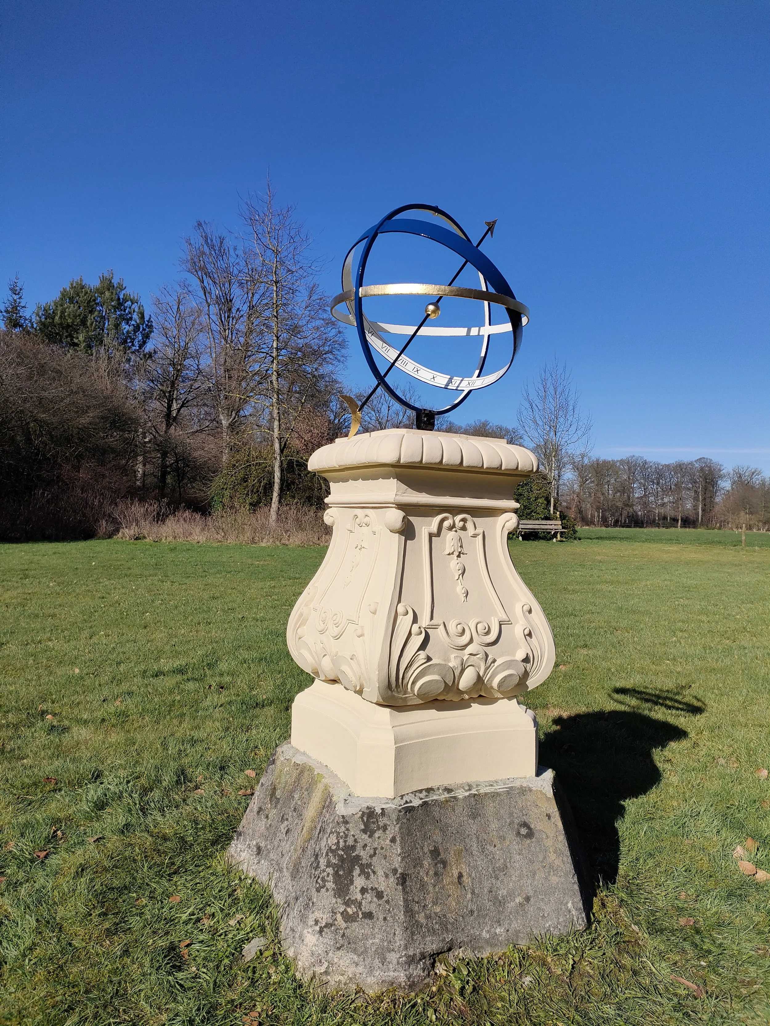 Een outdoor astronomisch uurwerk met een model van een planetenstelsel op een sierlijke voet, onder een heldere blauwe lucht, omgeven door bomen en gras.