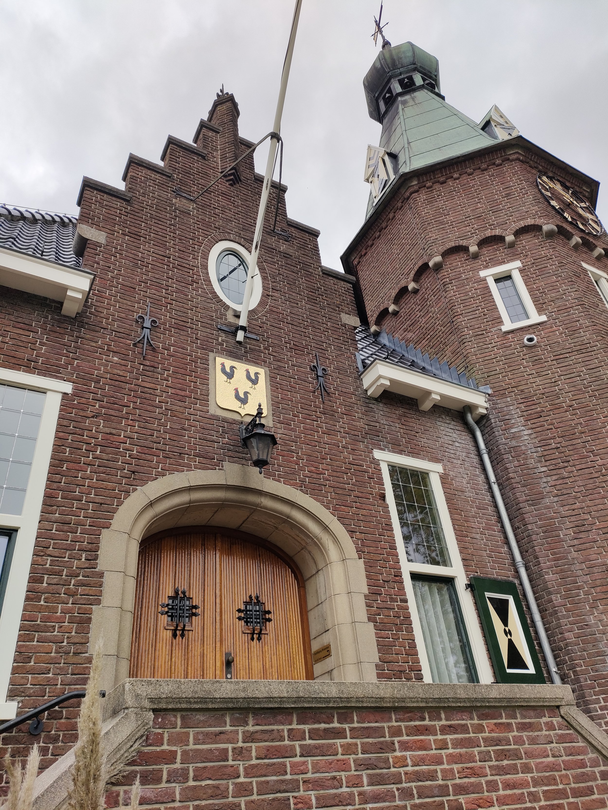 Een stenen poort met houten deur op een historische toren met een wijzerplaat en vlaggenmast, omgeven door een muur en ramen, typisch voor een oud kasteel of gemeentehuis.