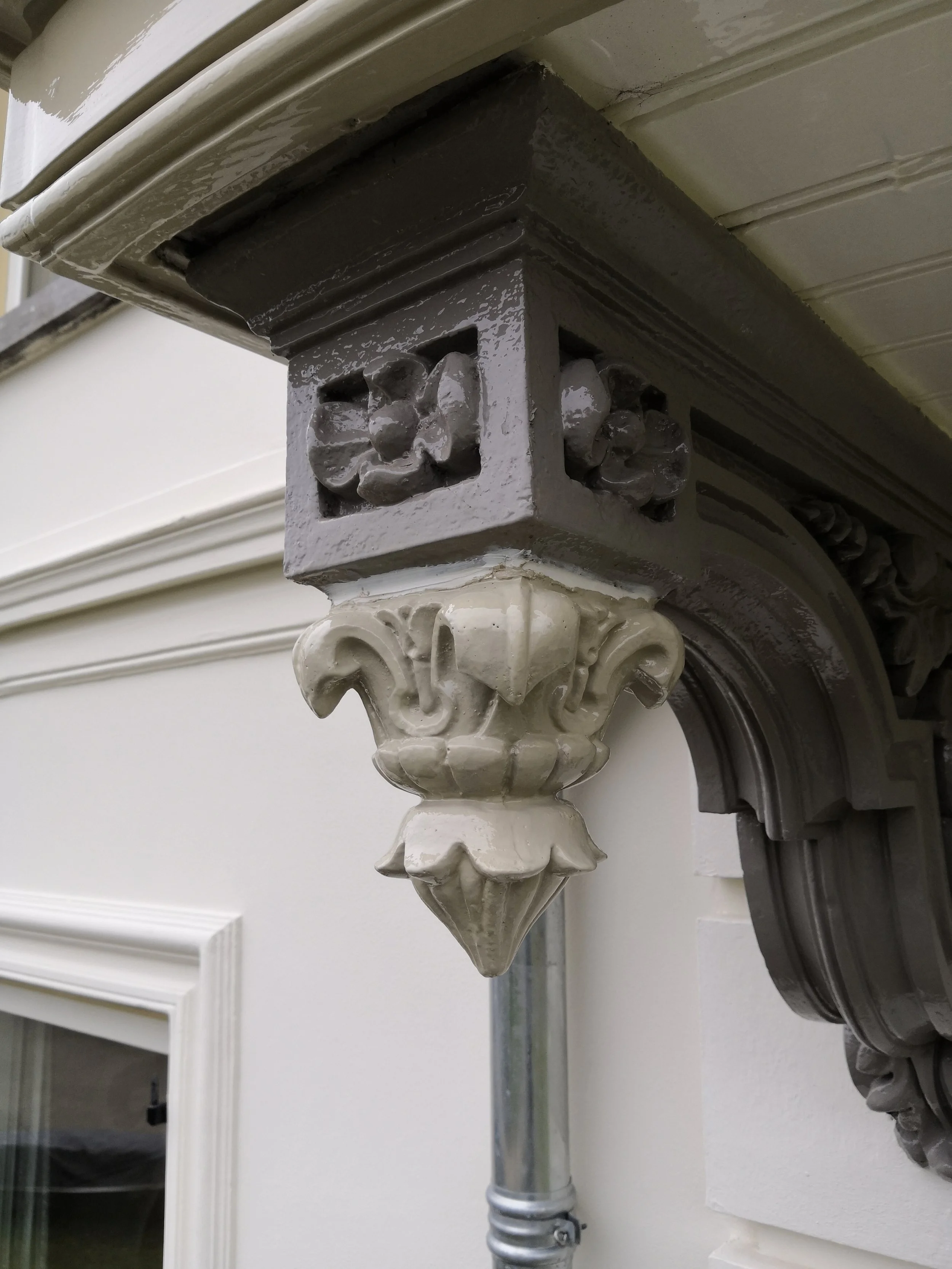 Details van een sierlijke muurdecoratie en een goot, bevestigd aan een witte muur bij een raam, met een metalen buis eronder.