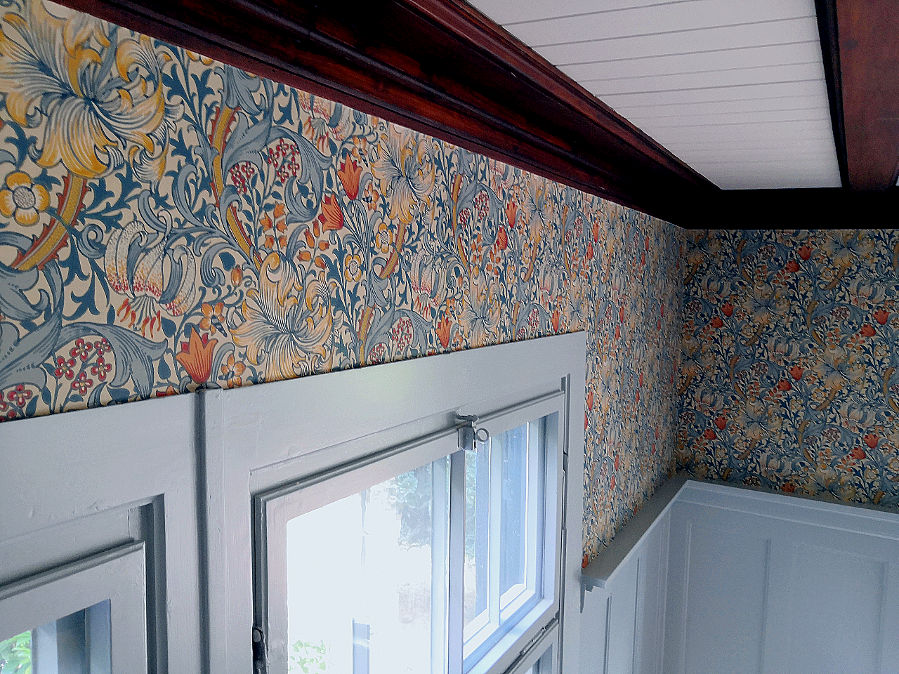 Een kamer met kleurrijke bloemenbehang en een witte raam met een gordijnrail ernaast.