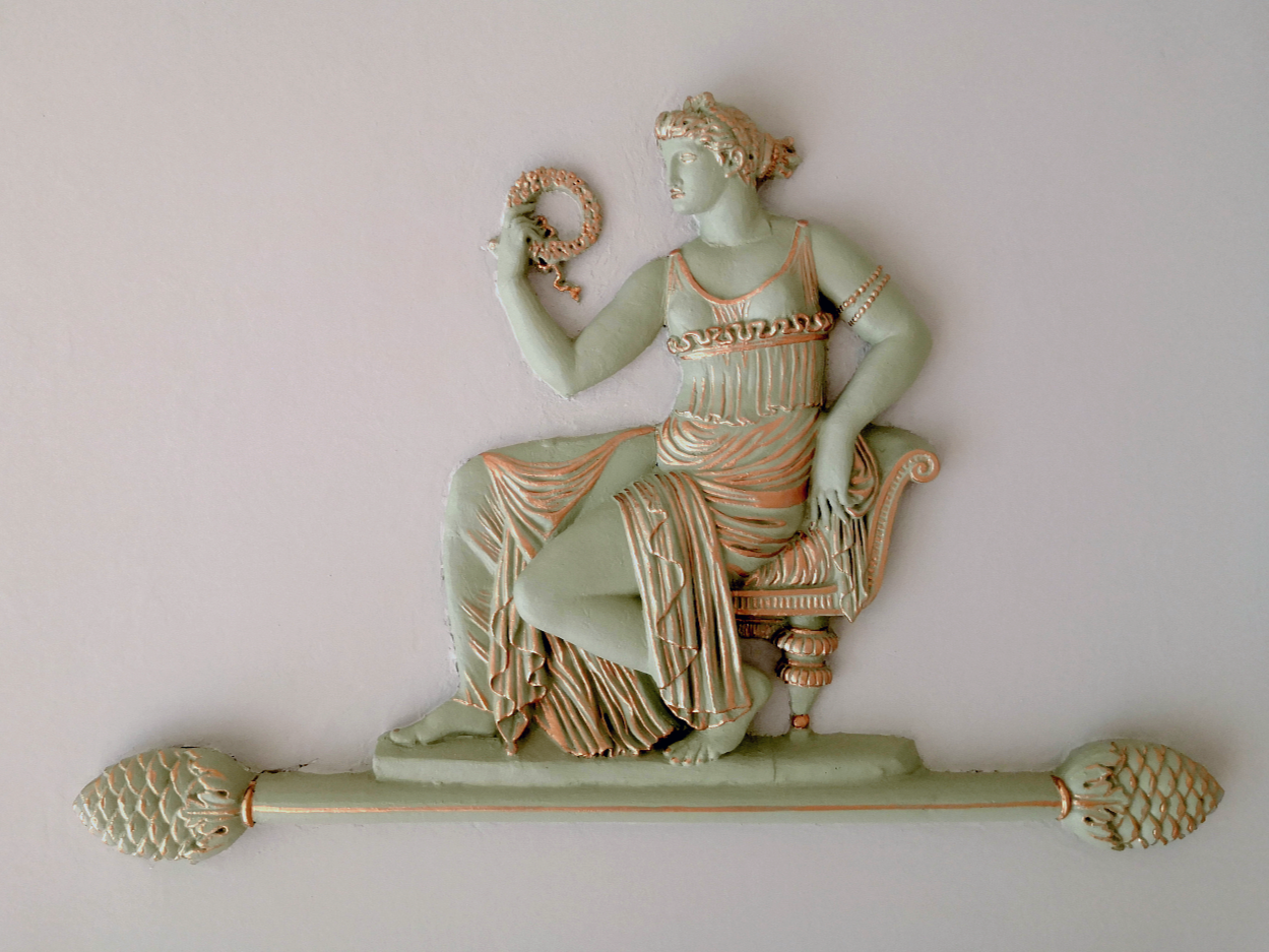 Een muursculptuur van een vrouw die op een stoel zit, met een staf en een slang in haar hand, in klassieke Griekse stijl.