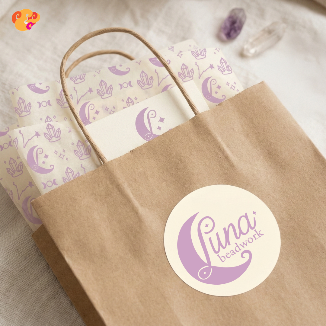 A brown paper shopping bag with a white circular logo reading 'Luna beadwork' in purple, sitting on a light-colored surface. Inside the bag, pastel purple-patterned tissue paper featuring moons, stars, and crystals is visible. Two crystals, one purple and one clear, rest nearby.