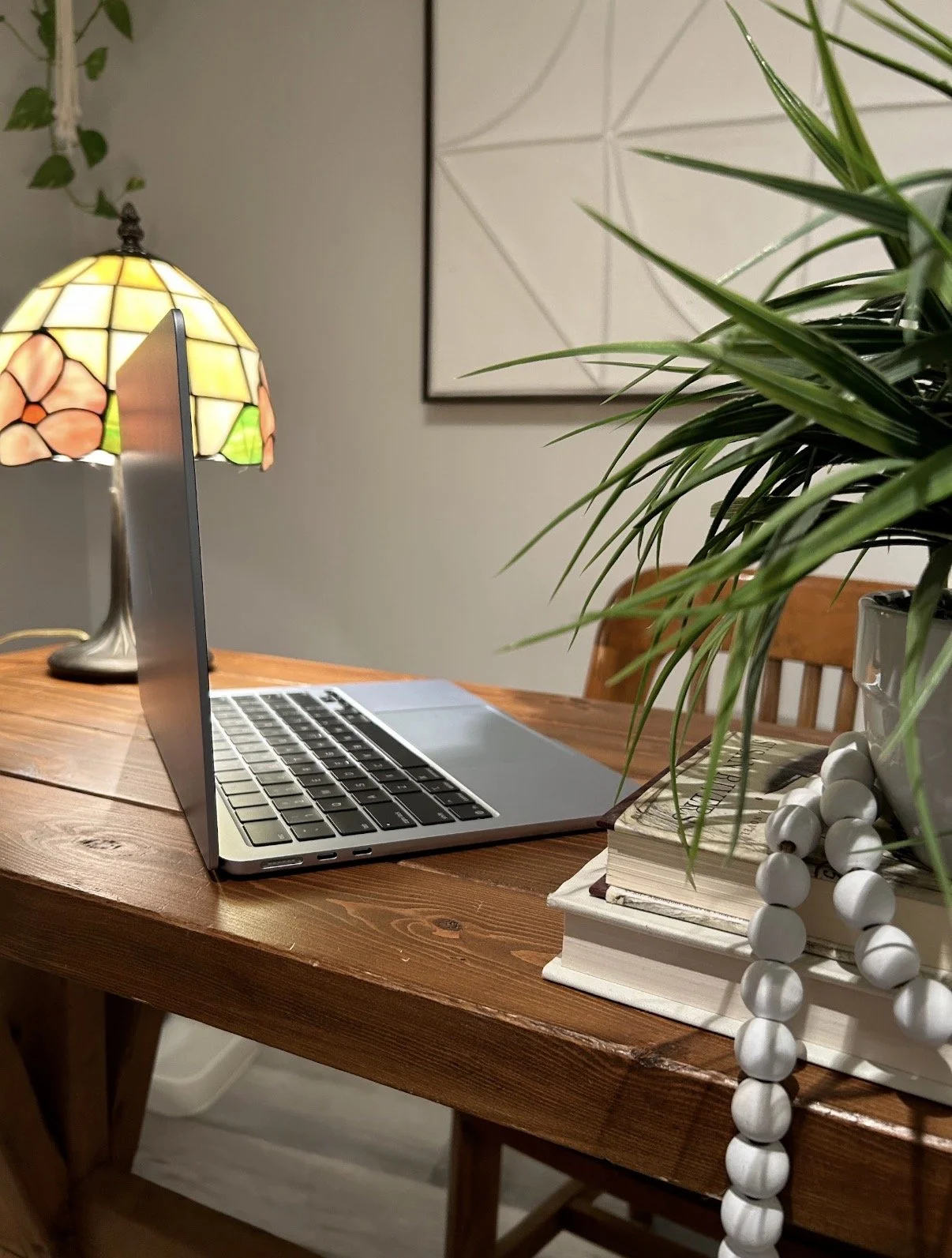 Wooden desk with a laptop, a stack of books, a potted plant with green leaves, and a Tiffany-style table lamp in a cozy room.