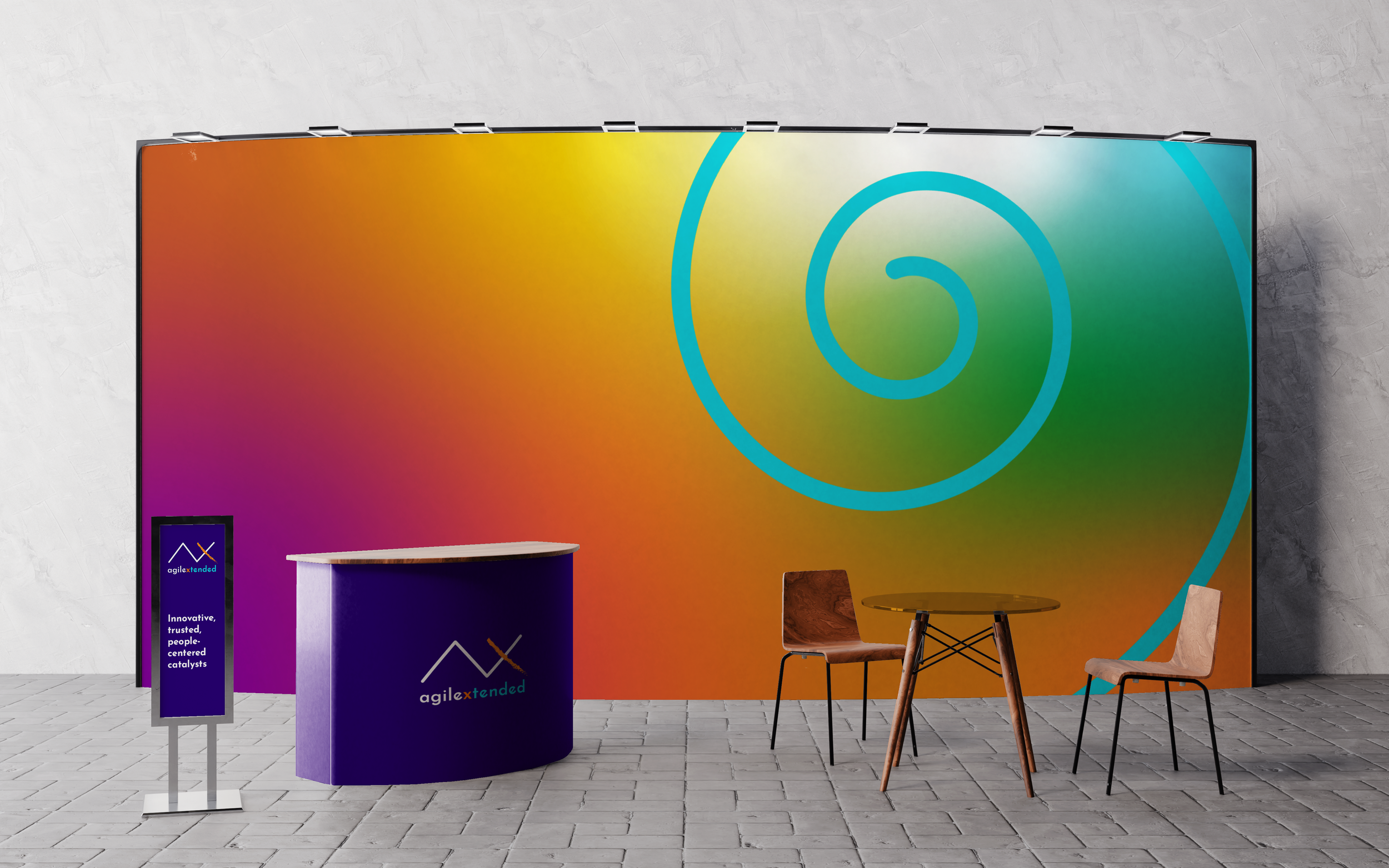 Exhibition booth with a purple curved counter with 'agilextended' logo, a purple standing sign with the same logo and the words 'Innovative, trusted, people-centered catalysts', a colorful large backdrop with a spiral design, two chairs, and a small round table.