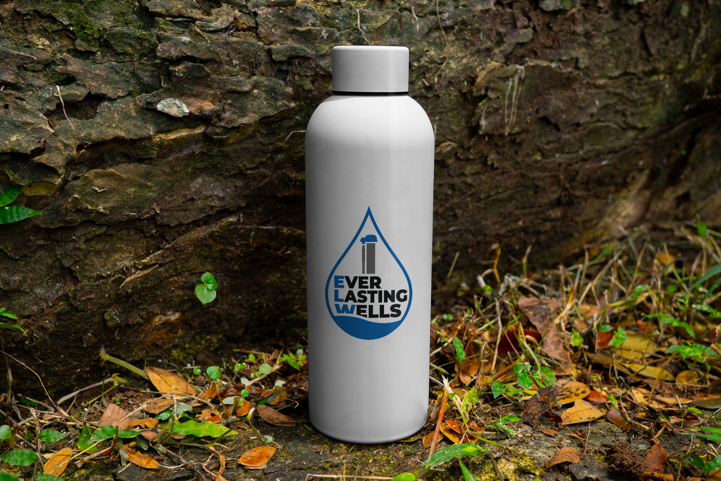 A white water bottle with a logo that says 'Everlasting Wells' and features a graphic of a well and water droplet, placed outdoors against a mossy rock and surrounded by green plants and fallen leaves.