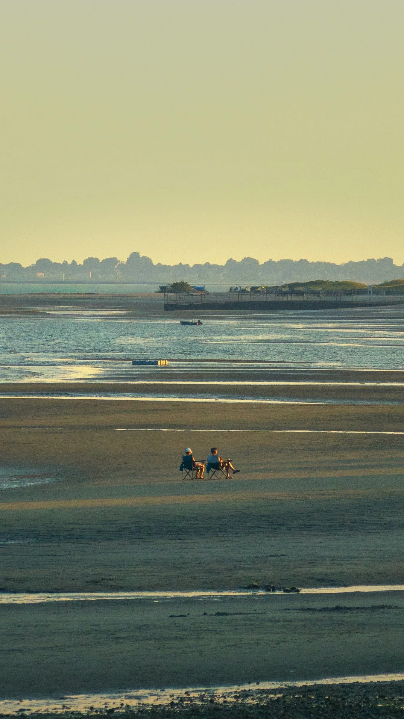 Two people sitting on camping chairs on a beach, looking towards the water with a boat in the distance.