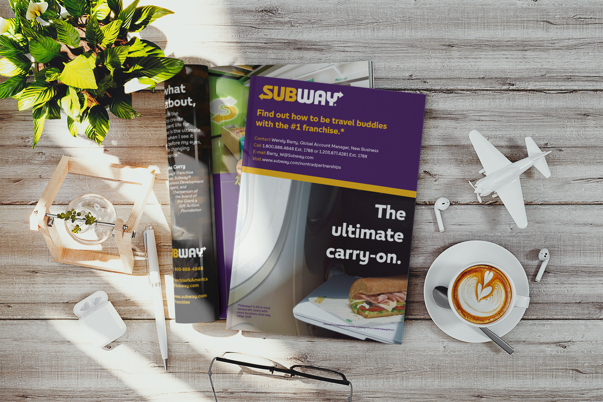 Flat lay of a wooden table with a Subway brochure, a white coffee cup with latte art, a small toy airplane, wireless earbuds, a pen, an open magazine, a pair of glasses, a white pen case, a small plant in a glass jar, and some sunlight.