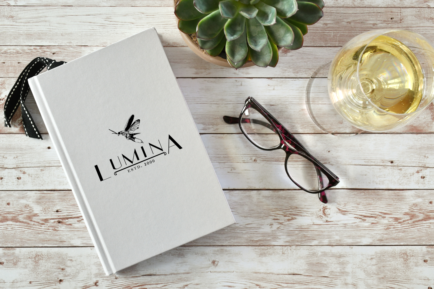 A white notebook with 'Lumina' and a dragonfly logo on a wooden table, next to glasses of white wine, a potted succulent plant, and a pair of black-rimmed glasses.