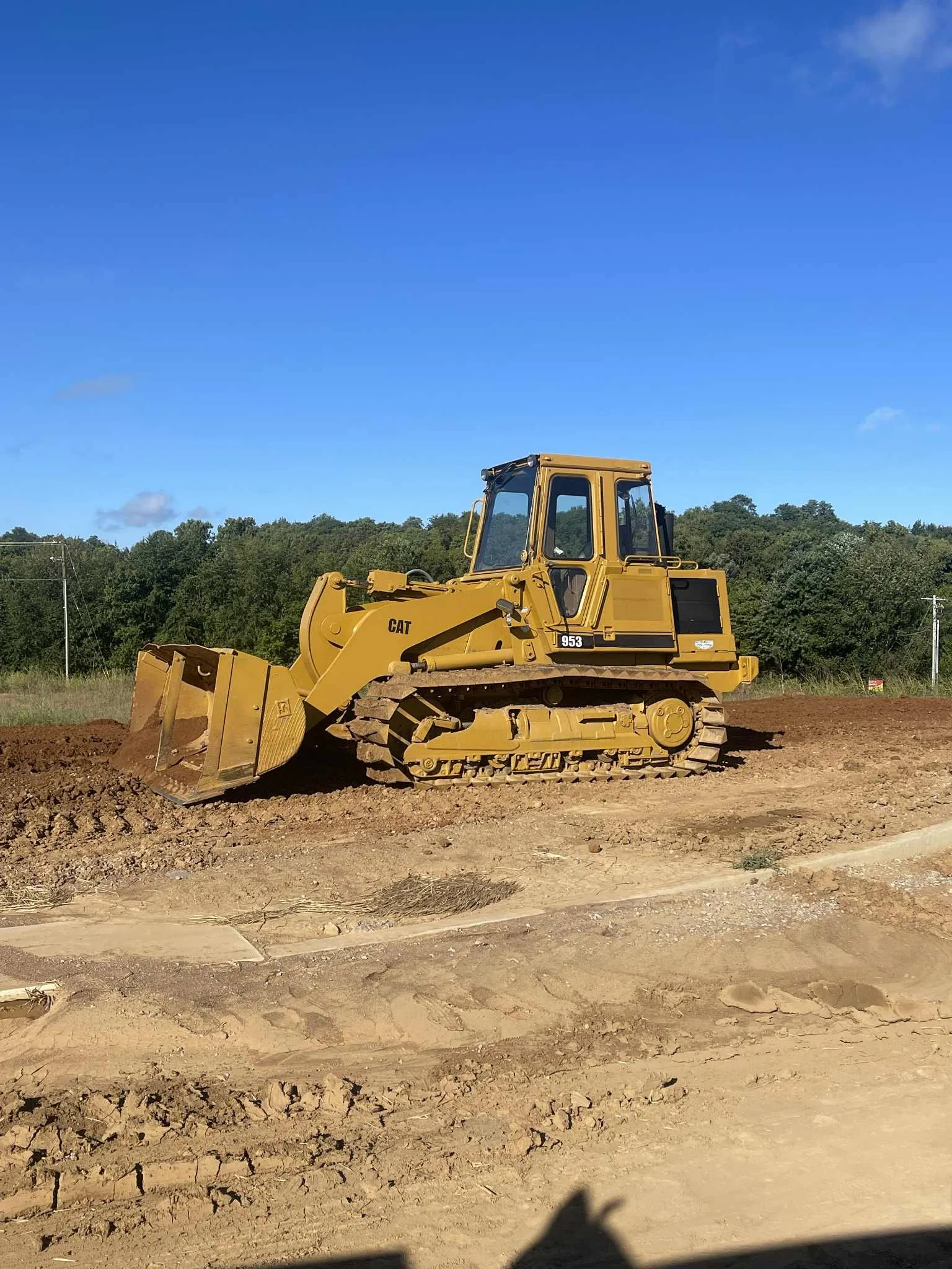 A yellow Caterpillar bulldozer on a construction site with dirt and cleared land, under a clear blue sky with some trees and power lines in the background.