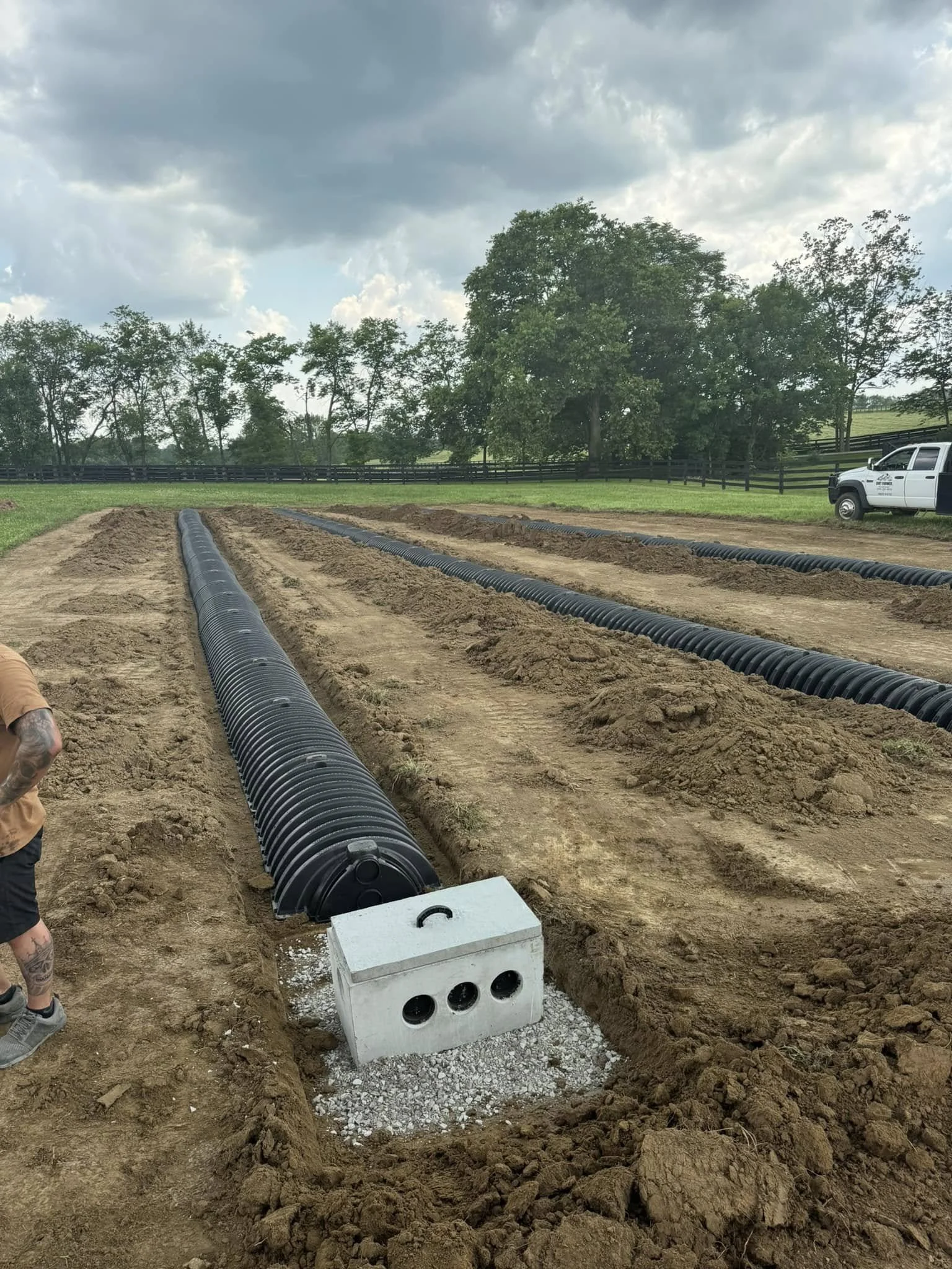 Construction site with black corrugated pipes laid in trenches on a farm, cloudy sky, green trees in the background, utility truck parked nearby, part of a tattooed person's arm visible.
