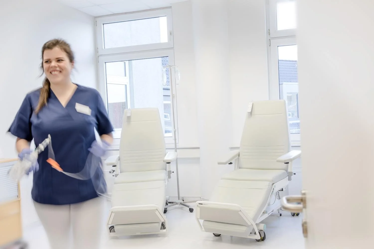 Eine Krankenschwester in blauer Uniform lächelt in einem Behandlungszimmer mit weißen medizinischen Stühlen und einem Infusionsständer vor einem Fenster.