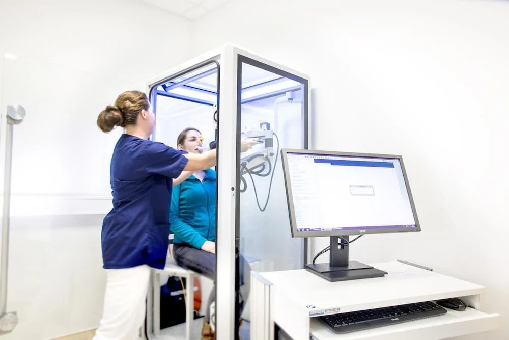 Eine medizinische Fachkraft führt eine Lungenfunktionstest an einer Patientin durch, die in einer Spirograph-Box sitzt.