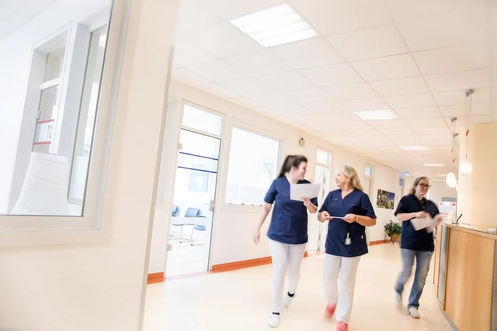 Drei Pflegekräfte in blauen Uniformen gehen in einem hell erleuchteten Krankenhausflur, ohne zu lächeln.